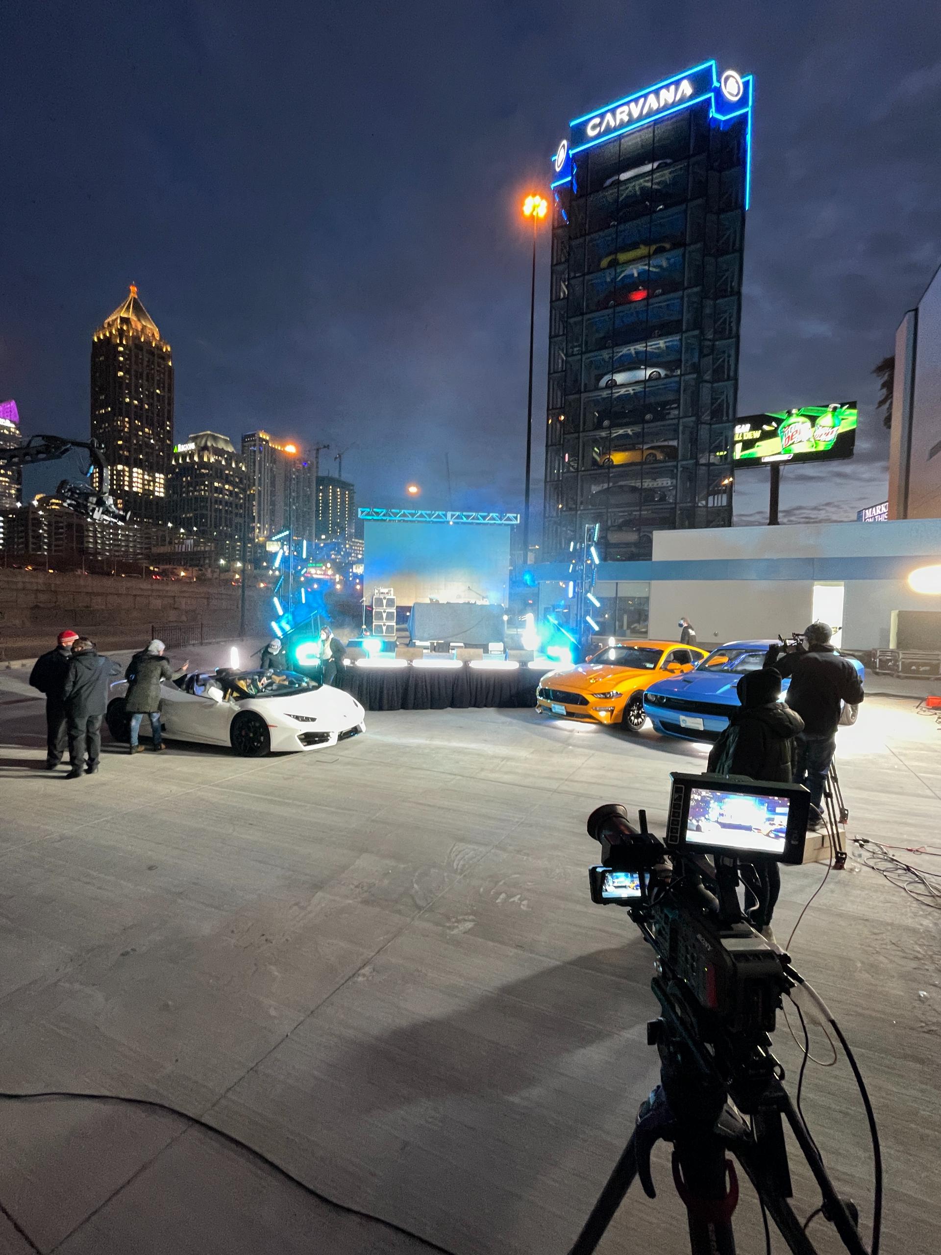 Outdoor nighttime TV production setup with cars and stage lighting.