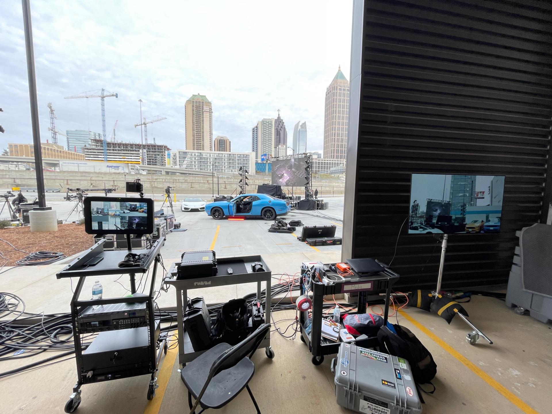 Behind-the-scenes view of a TV shoot with monitors, cables, and parked cars.