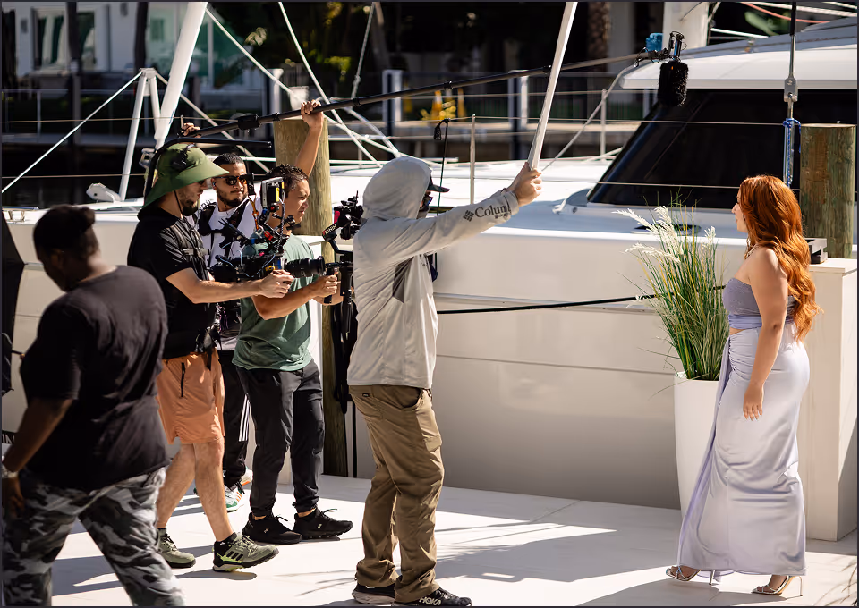 Crew filming TV commercial with boom mic and camera at marina, redheaded model in lavender dress on set.