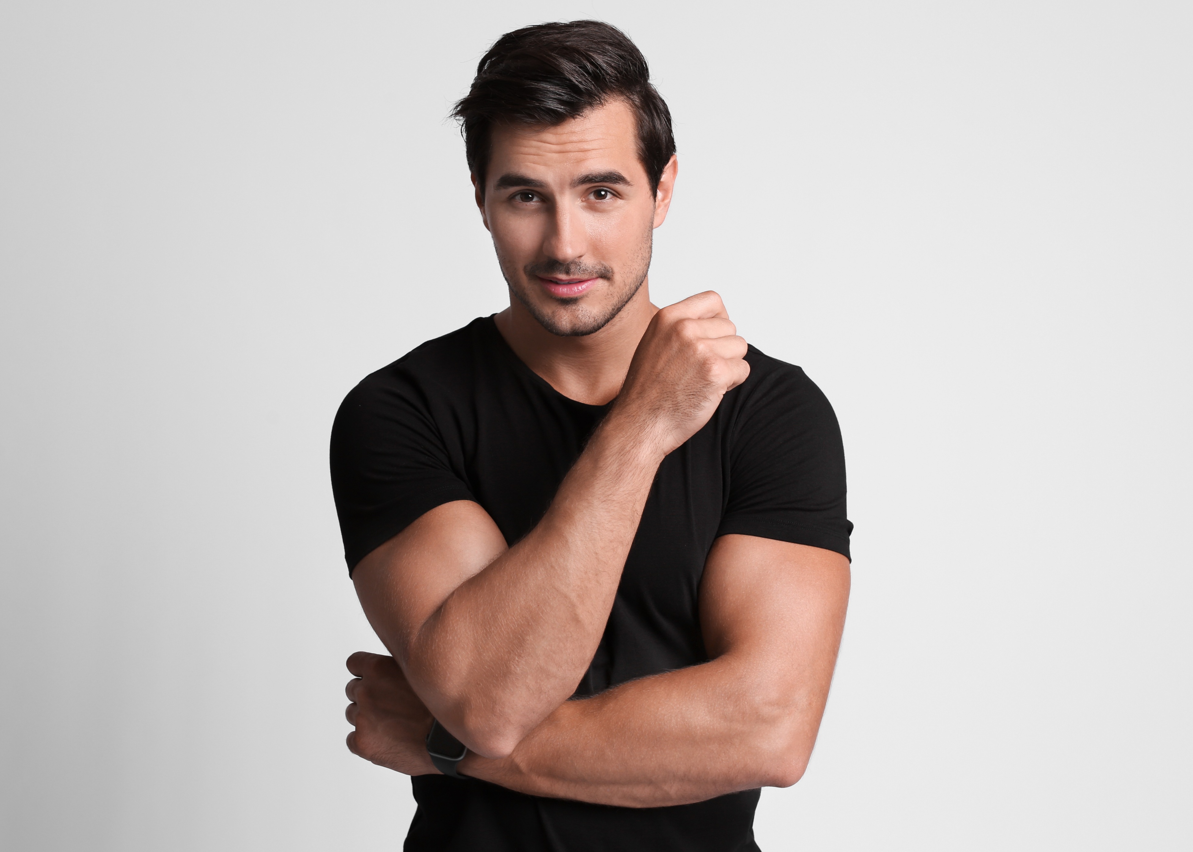 Man with dark hair wearing a black t-shirt, crossing his arms and looking confidently at the camera against a plain light gray background.