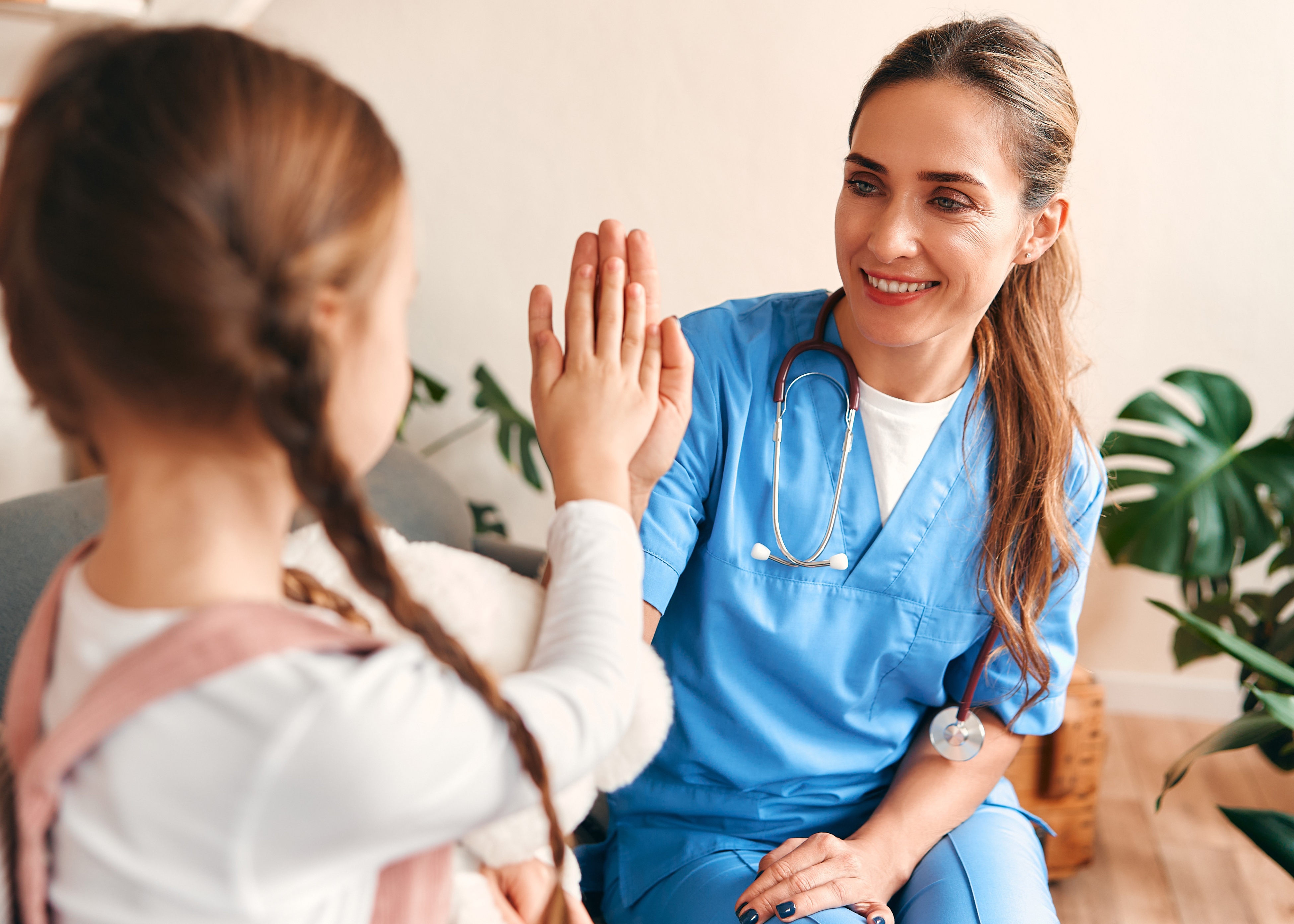 Female doctor in blue scrubs smiling and giving a high five to a young girl with braided hair.