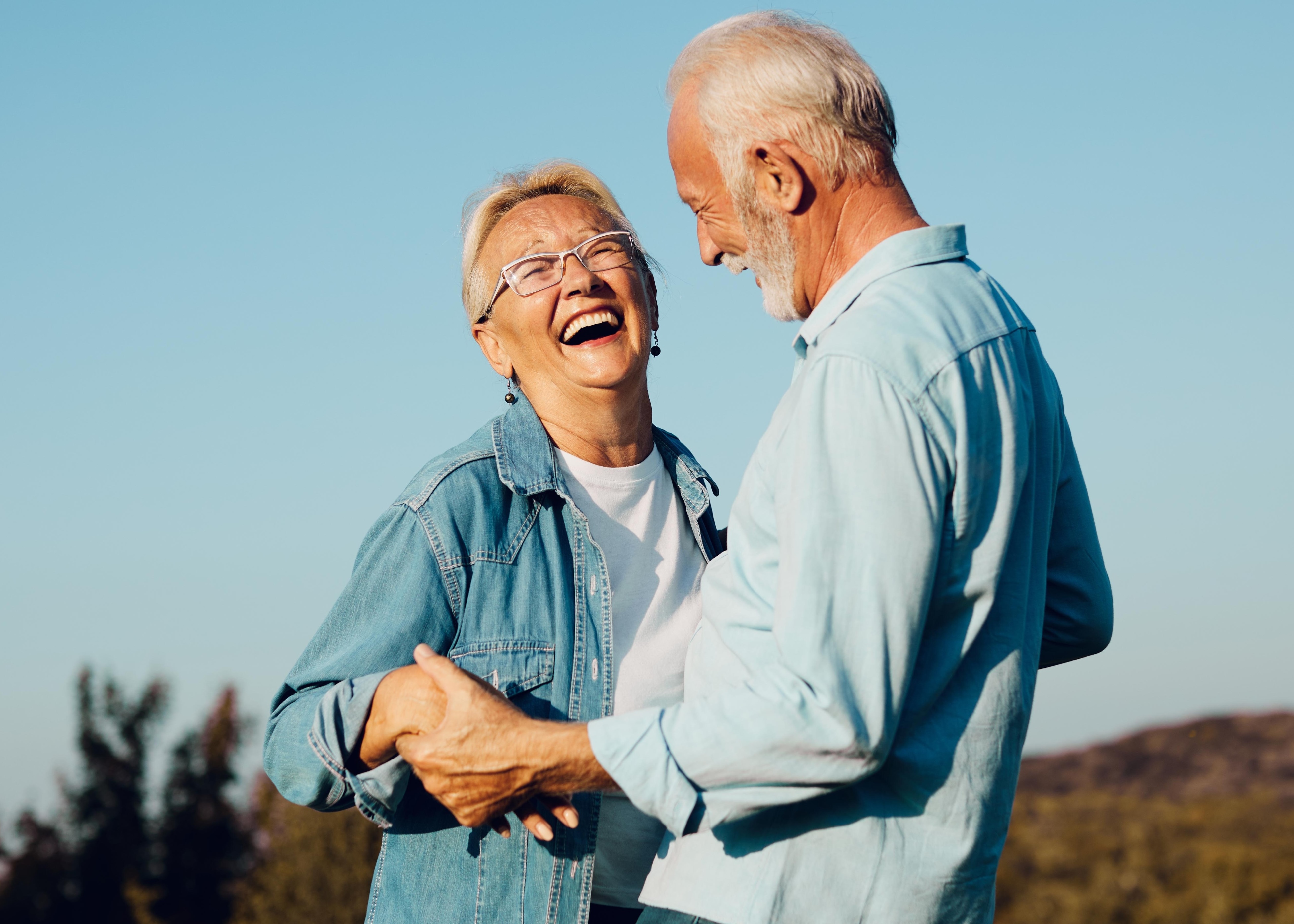 Elderly couple smiling and holding hands outdoors under a clear blue sky.