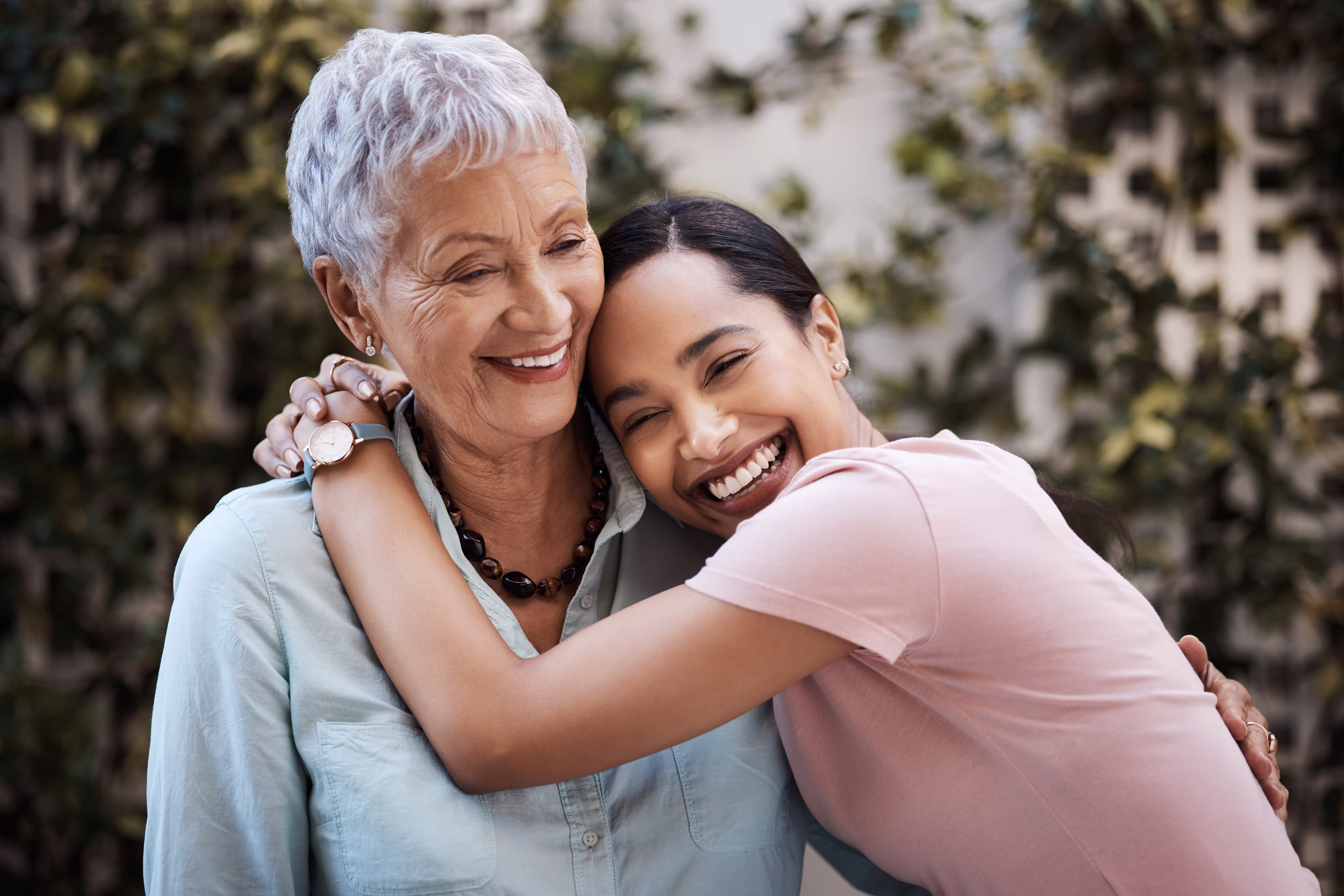 Young woman smiling and hugging an older woman outdoors with greenery in the background.