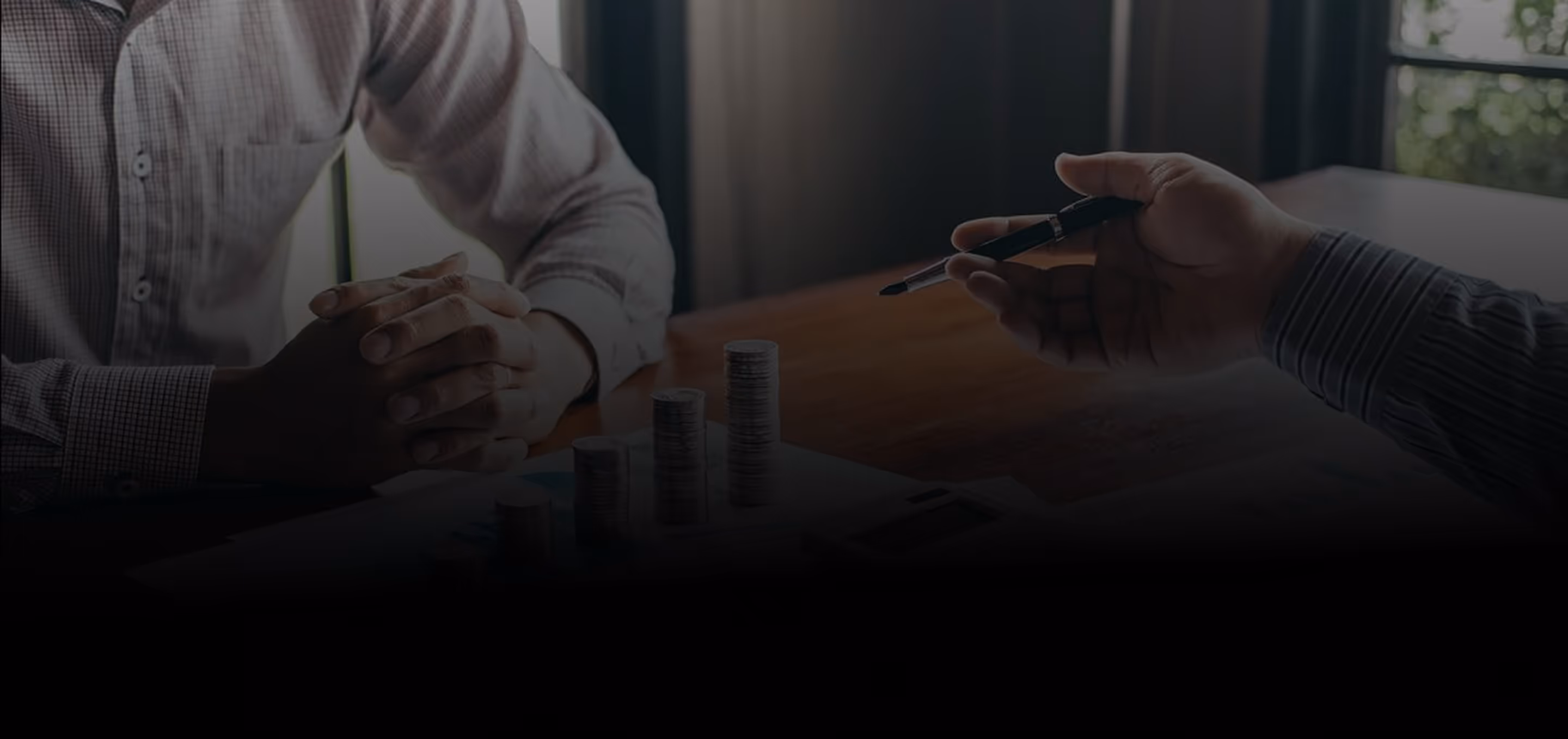Two people at a table with stacked coins, one holding a pen and the other with hands clasped, discussing finances.