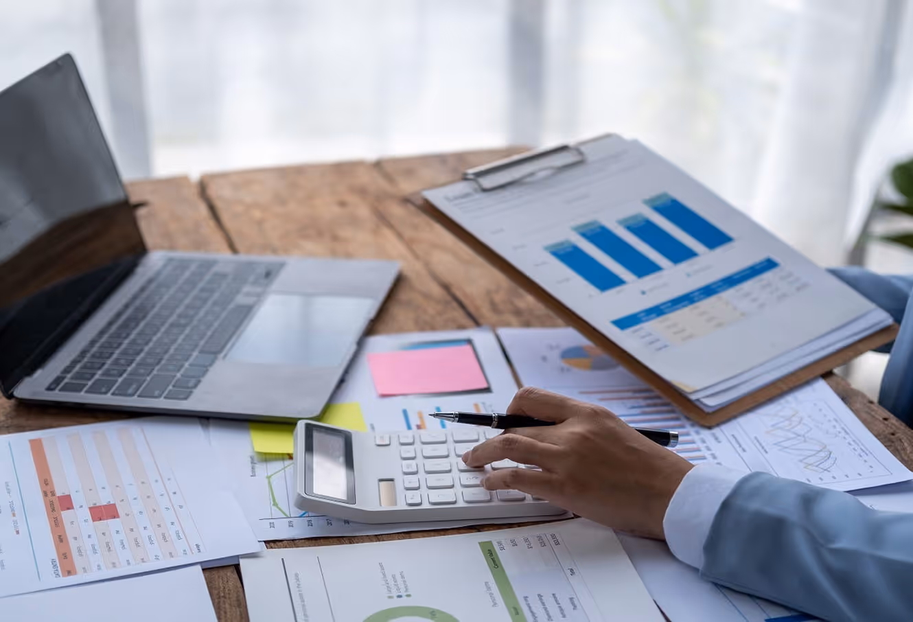 Person using a calculator and holding a pen with financial charts, graphs, and a laptop on a wooden desk.