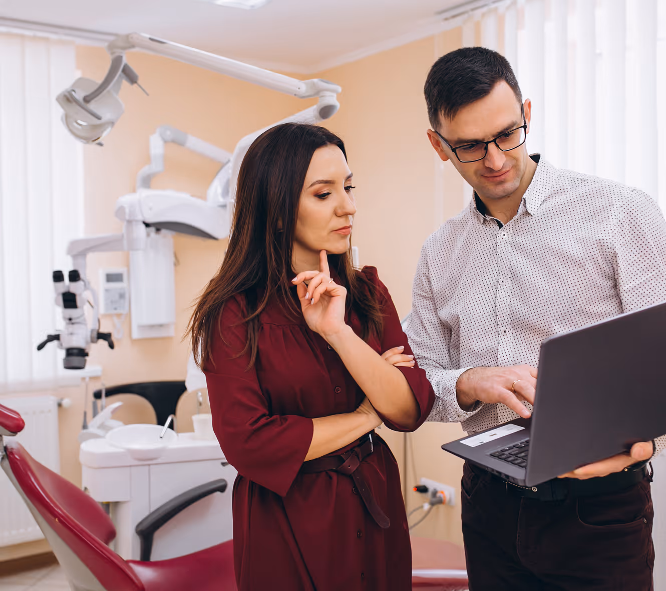 A man in glasses shows information on a laptop to a woman in a dental office with dental equipment and a maroon patient chair.