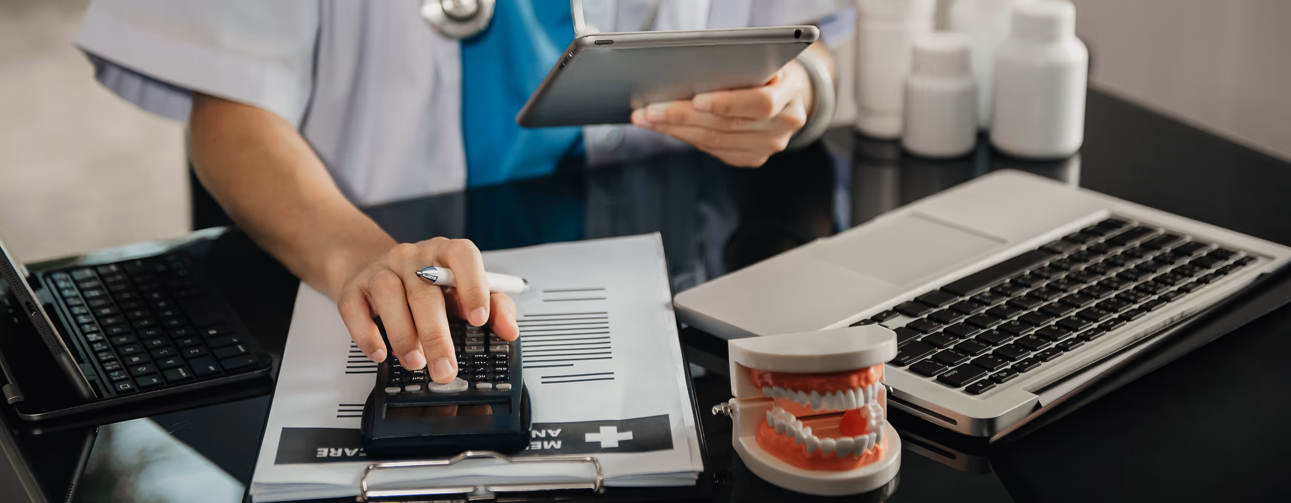 Medical professional using a calculator on a clipboard with medical documents, holding a tablet, next to a laptop and a dental model on a desk.