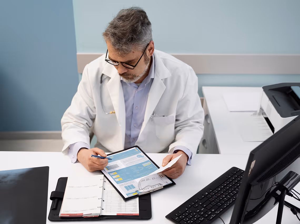 Middle-aged male doctor in a white coat reviewing medical documents at a desk with a computer and planner.