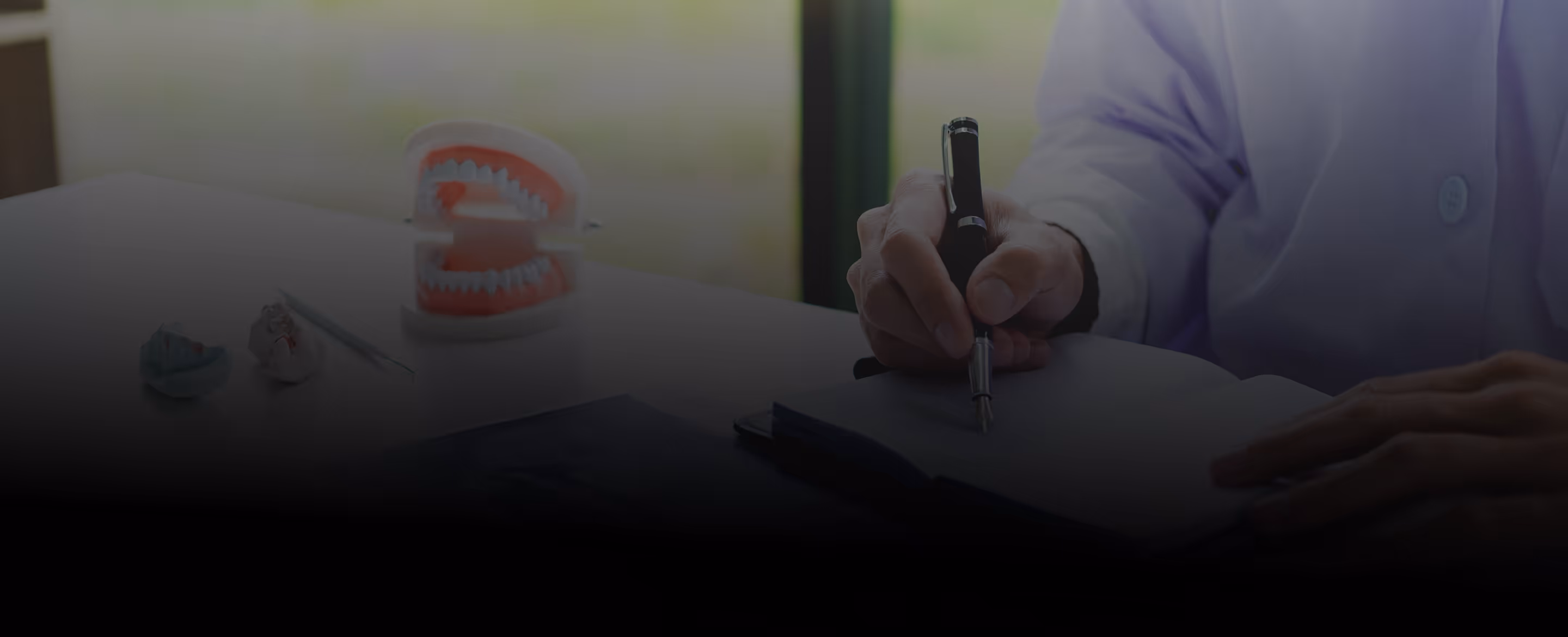Person in white coat writing with a pen at a desk with dental models and tools.