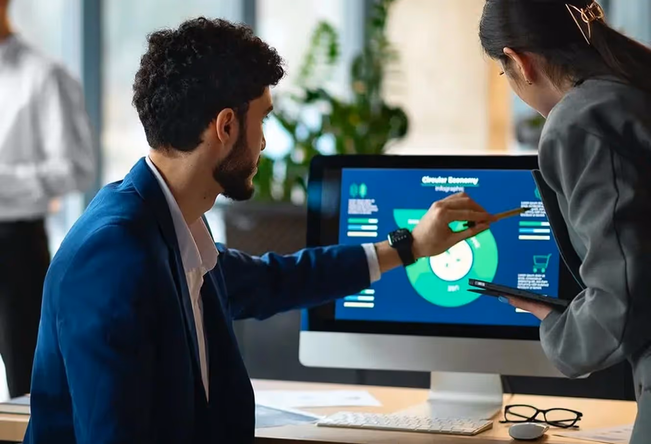 A man in a blue blazer points at a circular chart on a computer screen while a woman holds a tablet and listens.