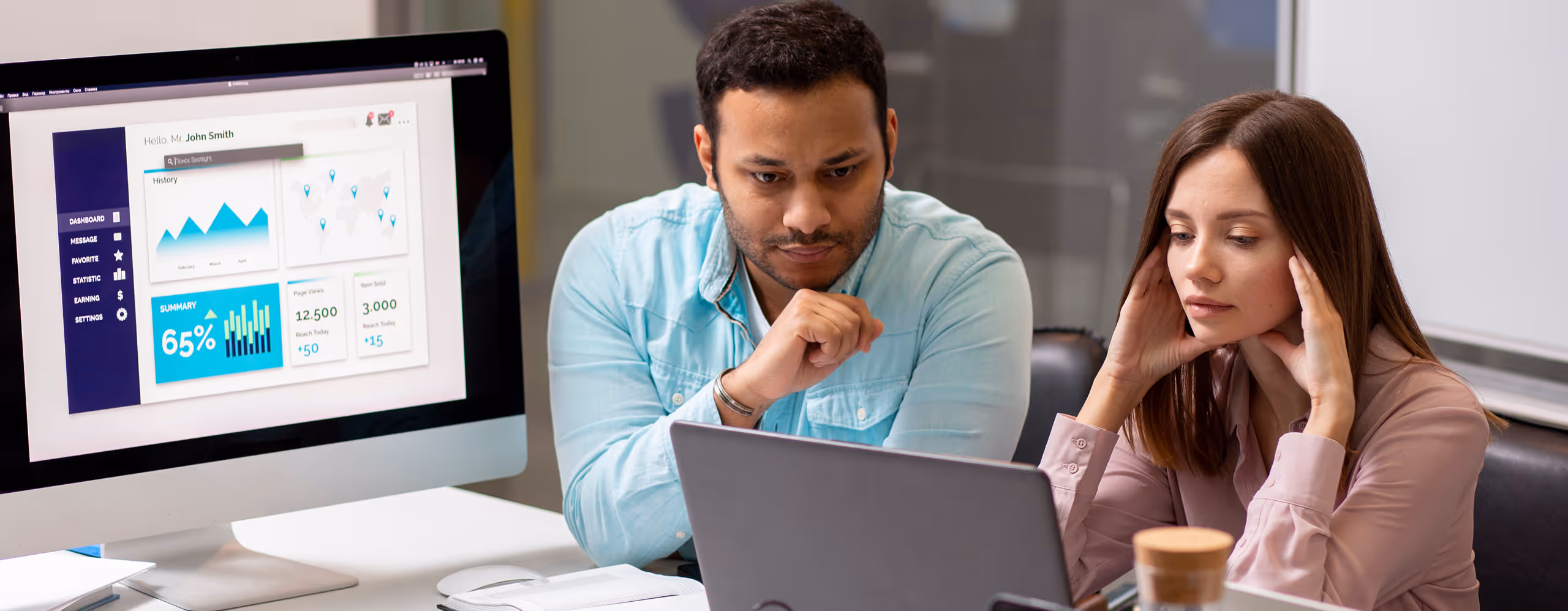 Two colleagues focused on a laptop screen in an office, with a desktop monitor displaying business analytics in the background.