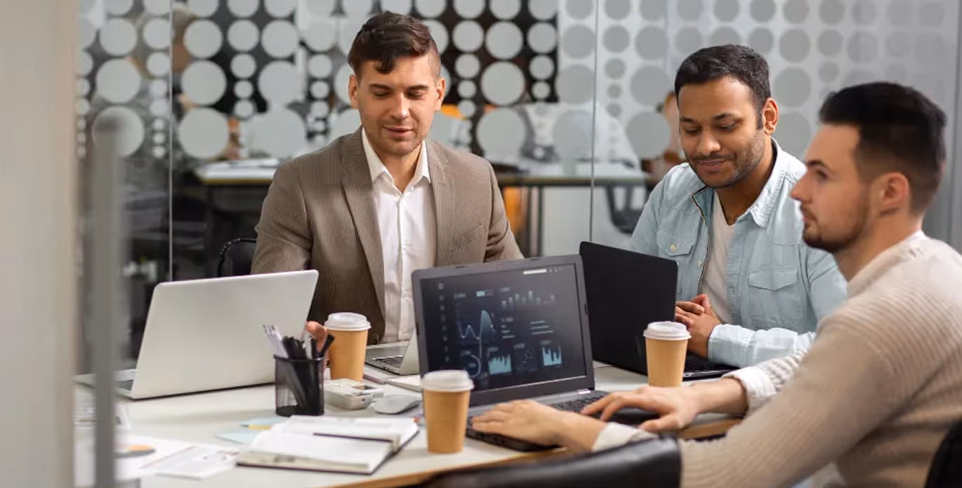 Three men working on laptops with coffee cups at a table in a modern office.