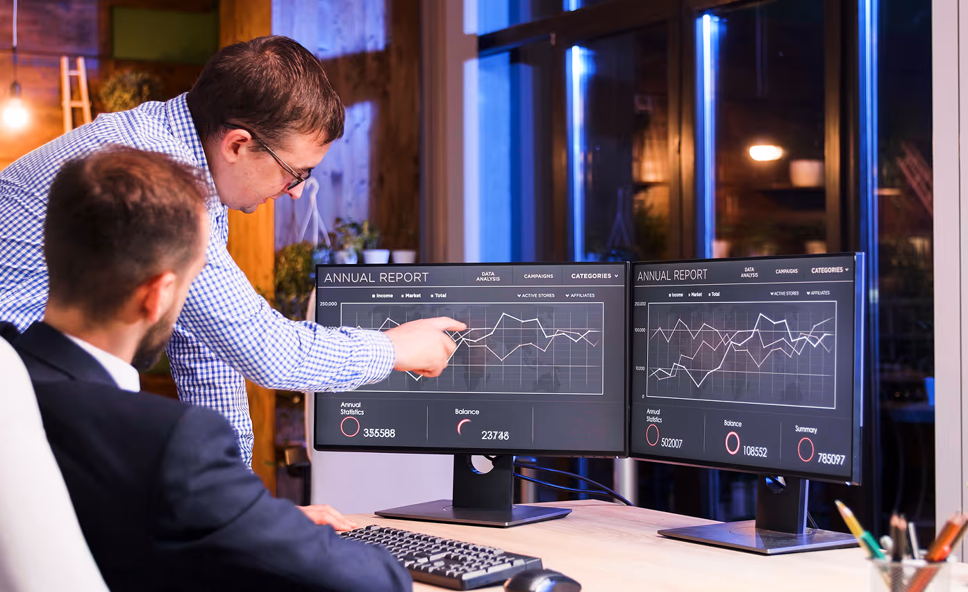 Two men discussing annual report graphs displayed on dual monitors in an office setting at night.