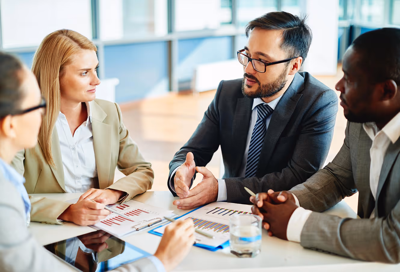 Four business professionals in a meeting discussing charts and graphs on paper and a tablet.