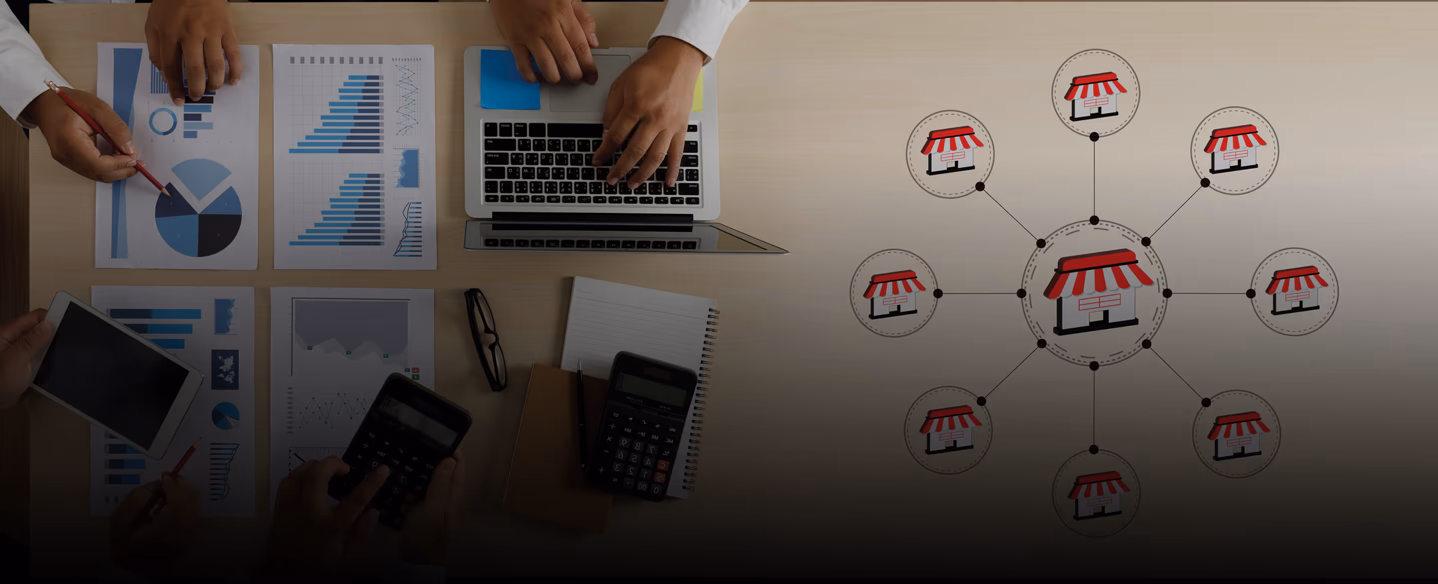 Overhead view of a desk with hands working on charts, a laptop, tablet, calculators, and a diagram of connected storefront icons representing a business network.