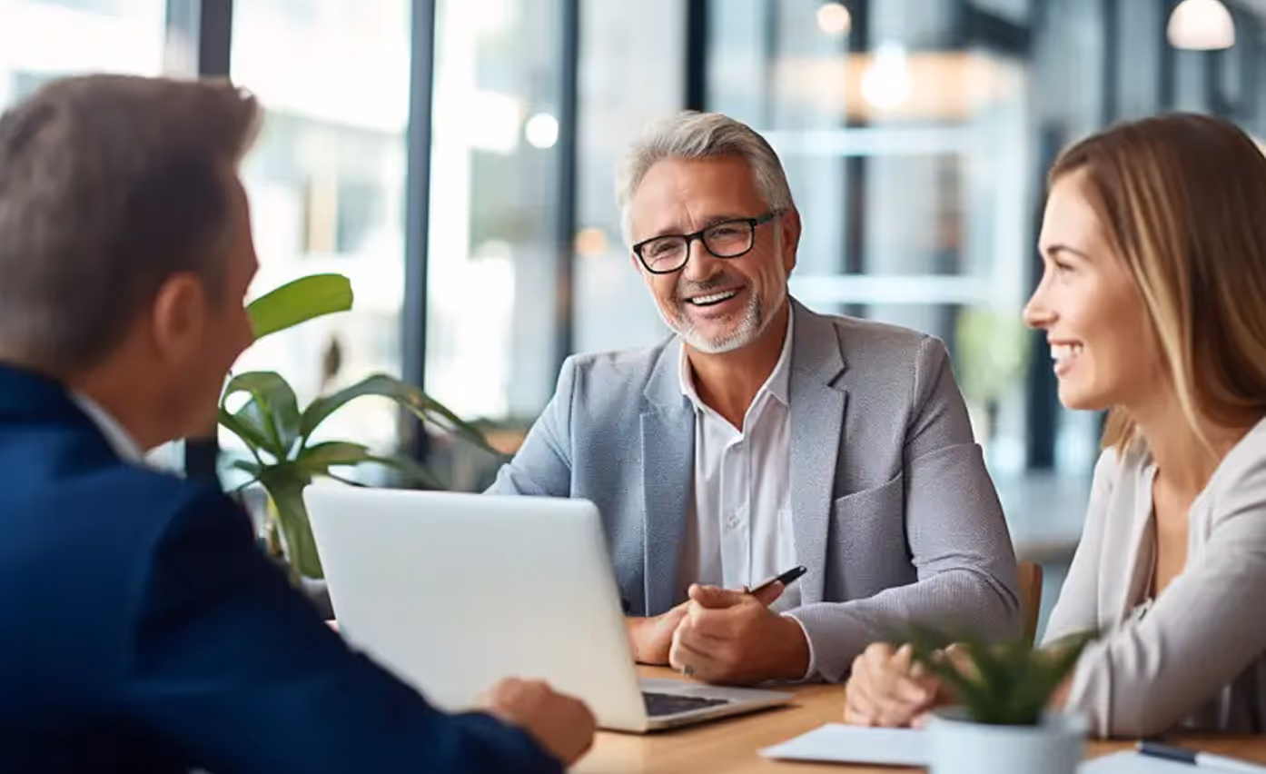 Three business professionals smiling and talking around a table with a laptop in a bright modern office.