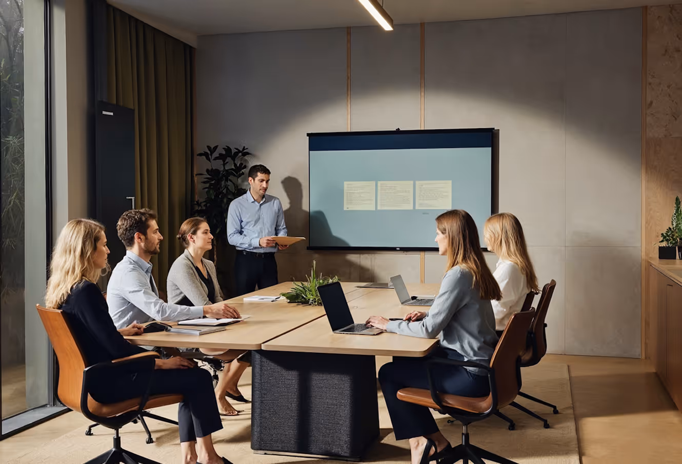 Business meeting with five colleagues seated around a conference table and one man standing presenting in front of a screen.