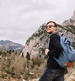 Man wearing sunglasses and a backpack hiking in a mountainous forested area.