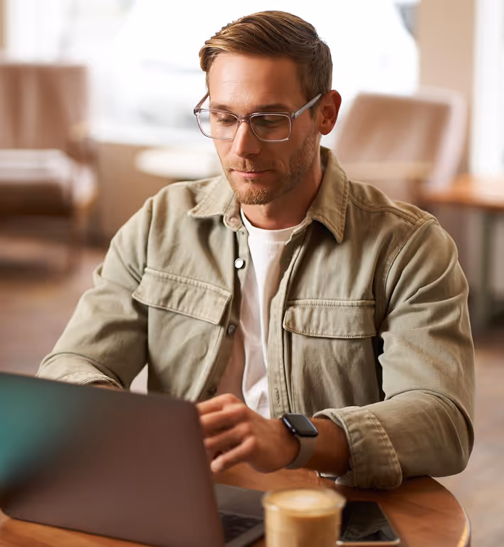 Man wearing glasses and a beige jacket working on a laptop at a round wooden table with a cup of coffee in a cozy room.