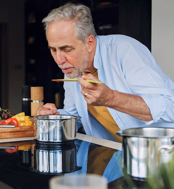 Older man smelling food from a spoon while cooking in a modern kitchen.