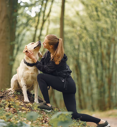 Woman in black athletic wear kneeling and affectionately holding the face of a sitting yellow Labrador dog in a wooded area.