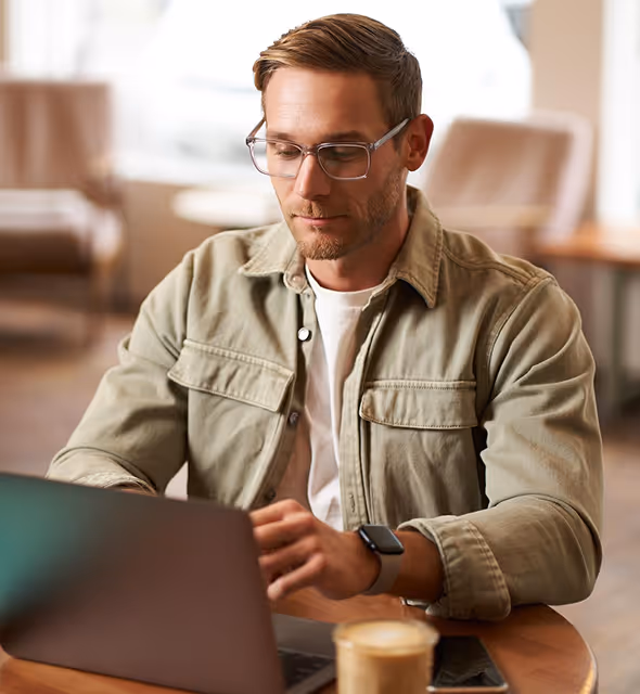 Young man with dark hair and beard wearing a black hoodie, focused on working on a silver laptop at a white desk indoors.