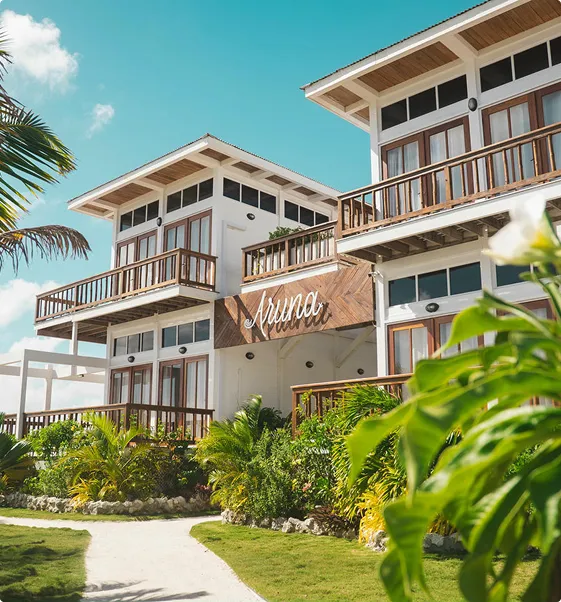 Two-story white building with wooden balconies and a sign reading 'Aruna' surrounded by tropical plants under a clear blue sky.