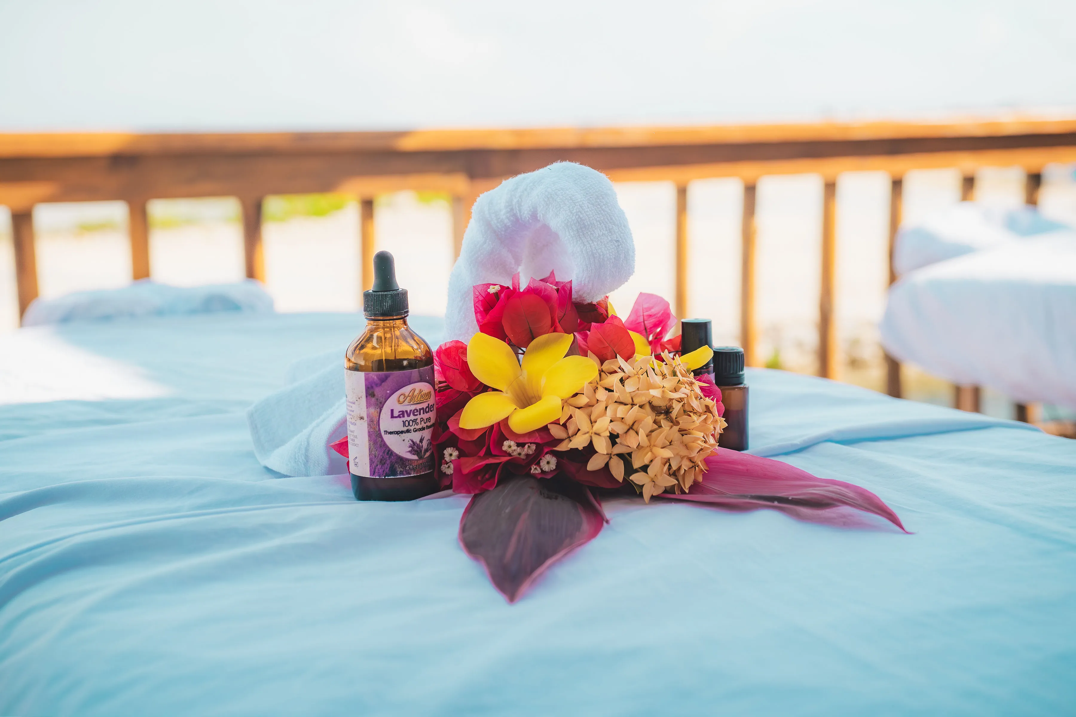 A lavender essential oil bottle, colorful flowers, and a rolled white towel on a blue spa table with wooden railing in the background.