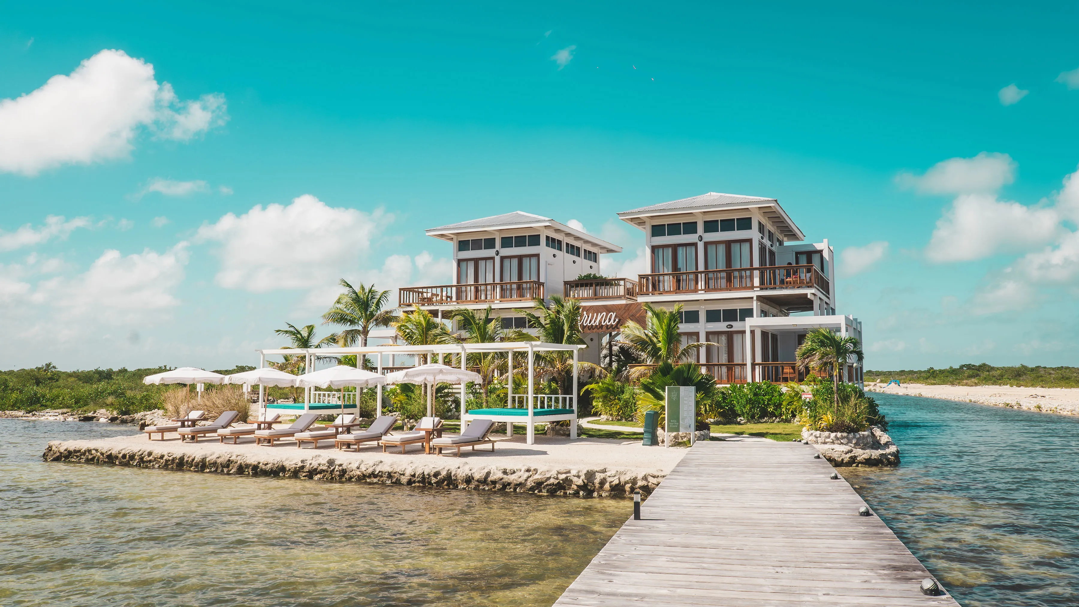 Modern white beachfront villa with two levels, surrounded by palm trees, sun loungers, umbrellas, and a wooden dock extending into clear water.