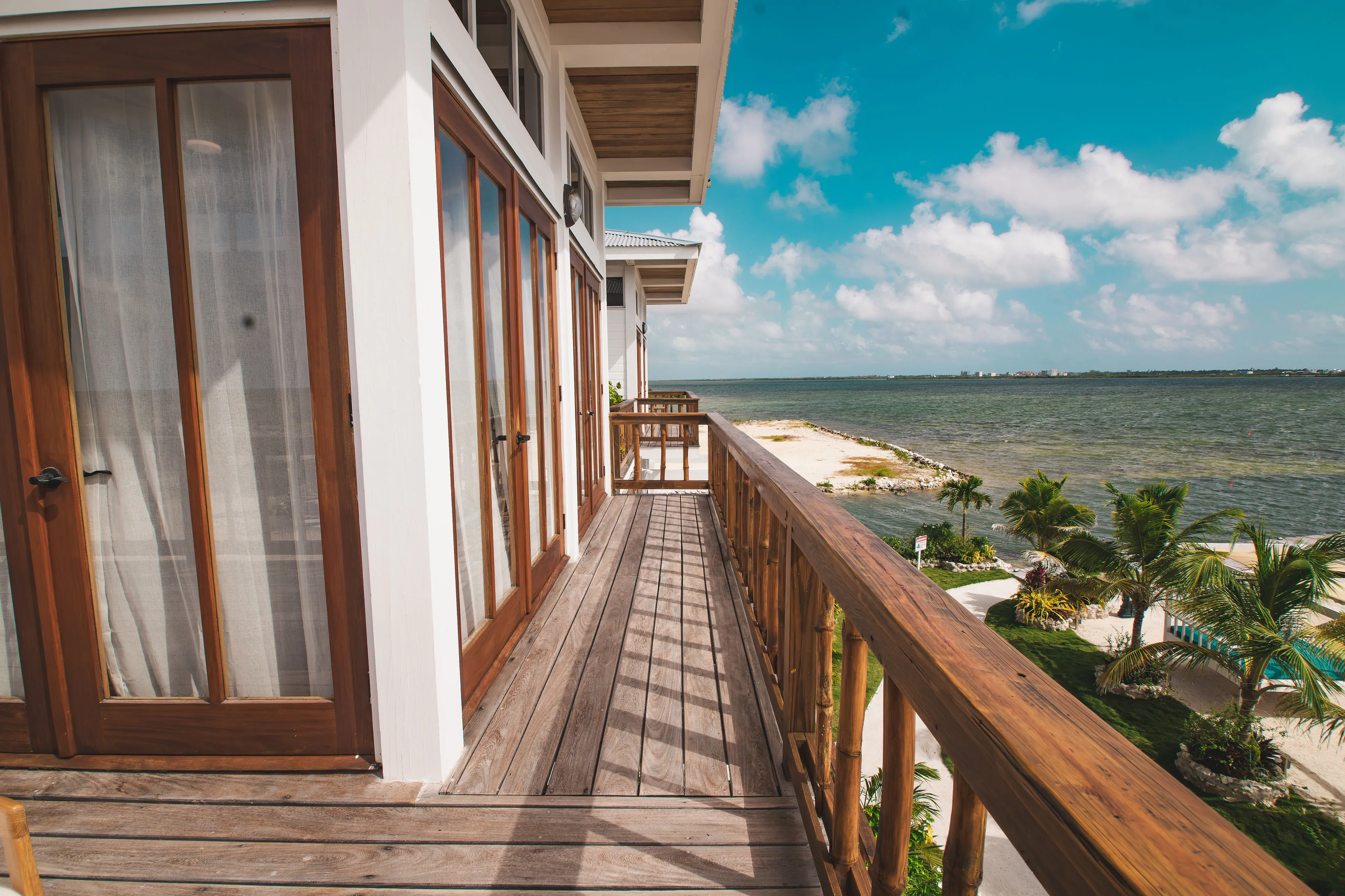 Wooden balcony with glass doors overlooking a beach and ocean under a partly cloudy sky.