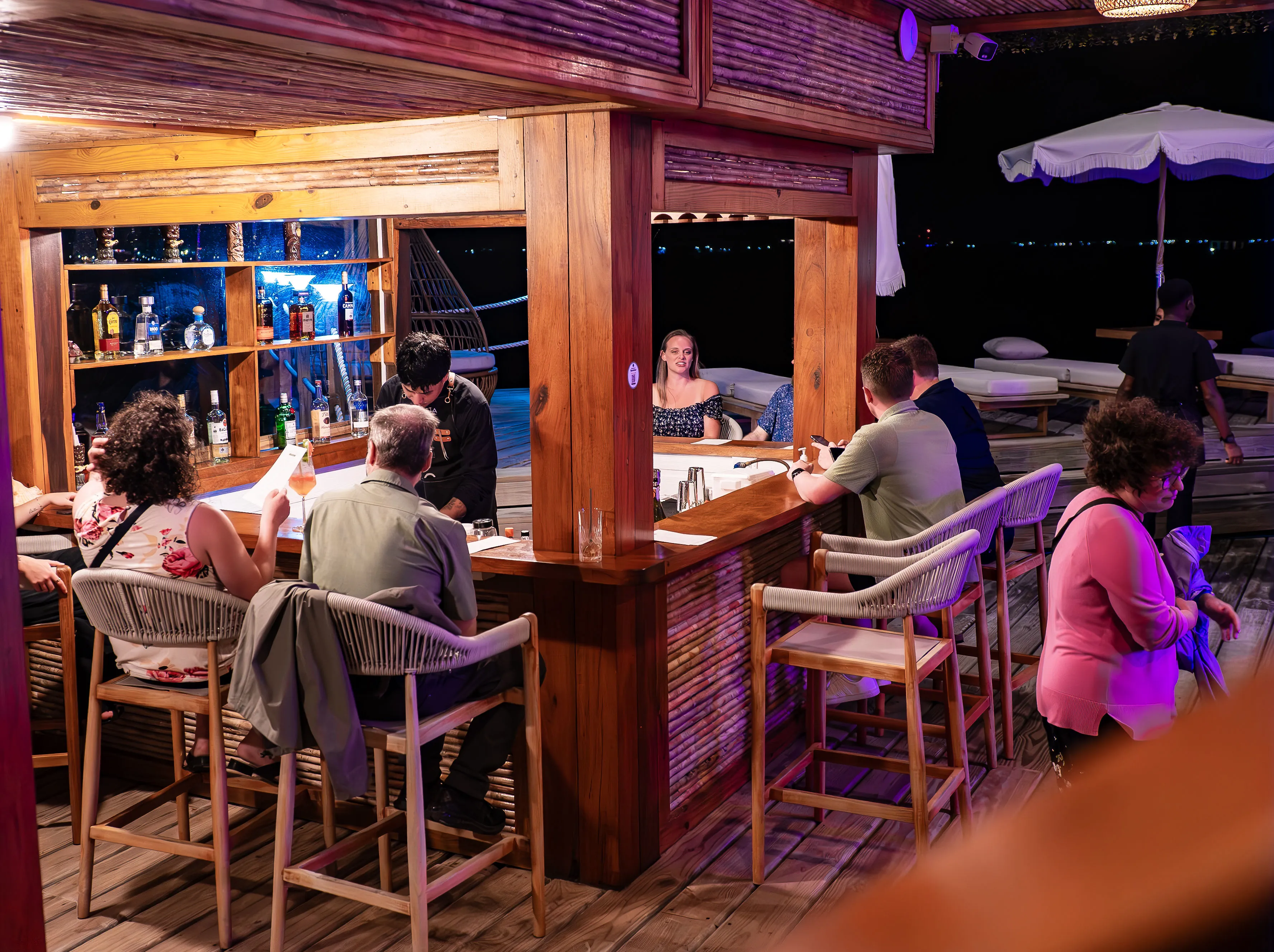 People seated at an outdoor wooden bar at night with bottles displayed on shelves and a bartender serving drinks.