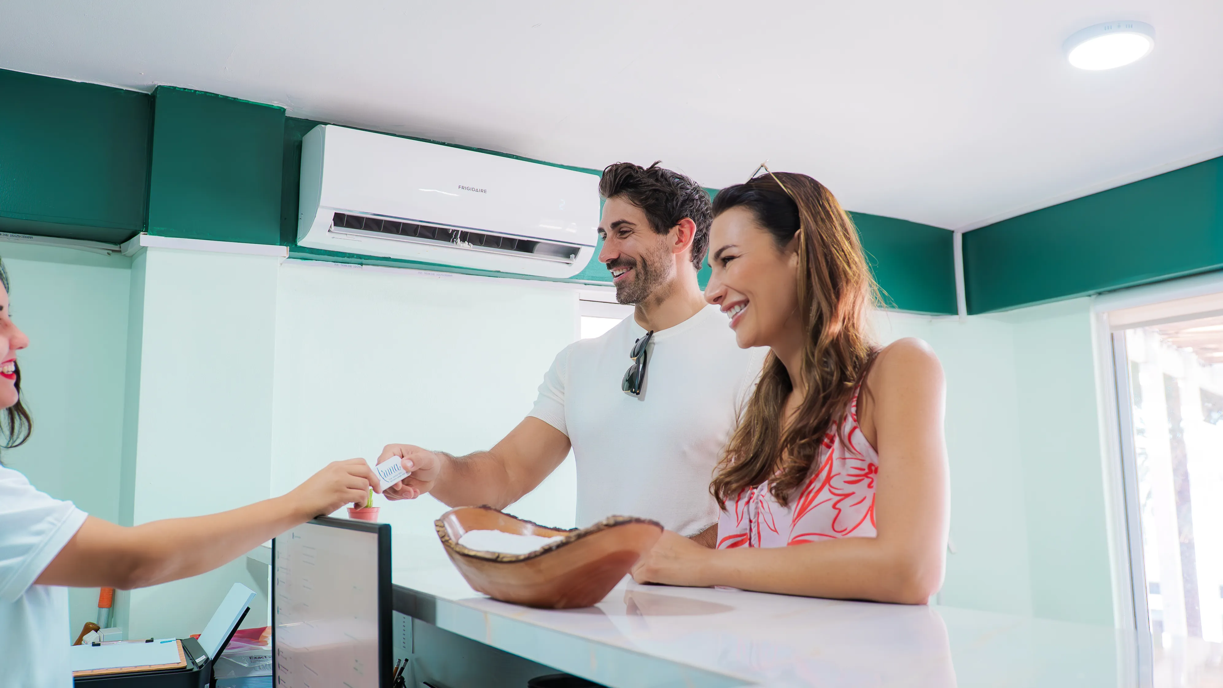A smiling couple handing a card to a receptionist at a bright, modern reception desk.