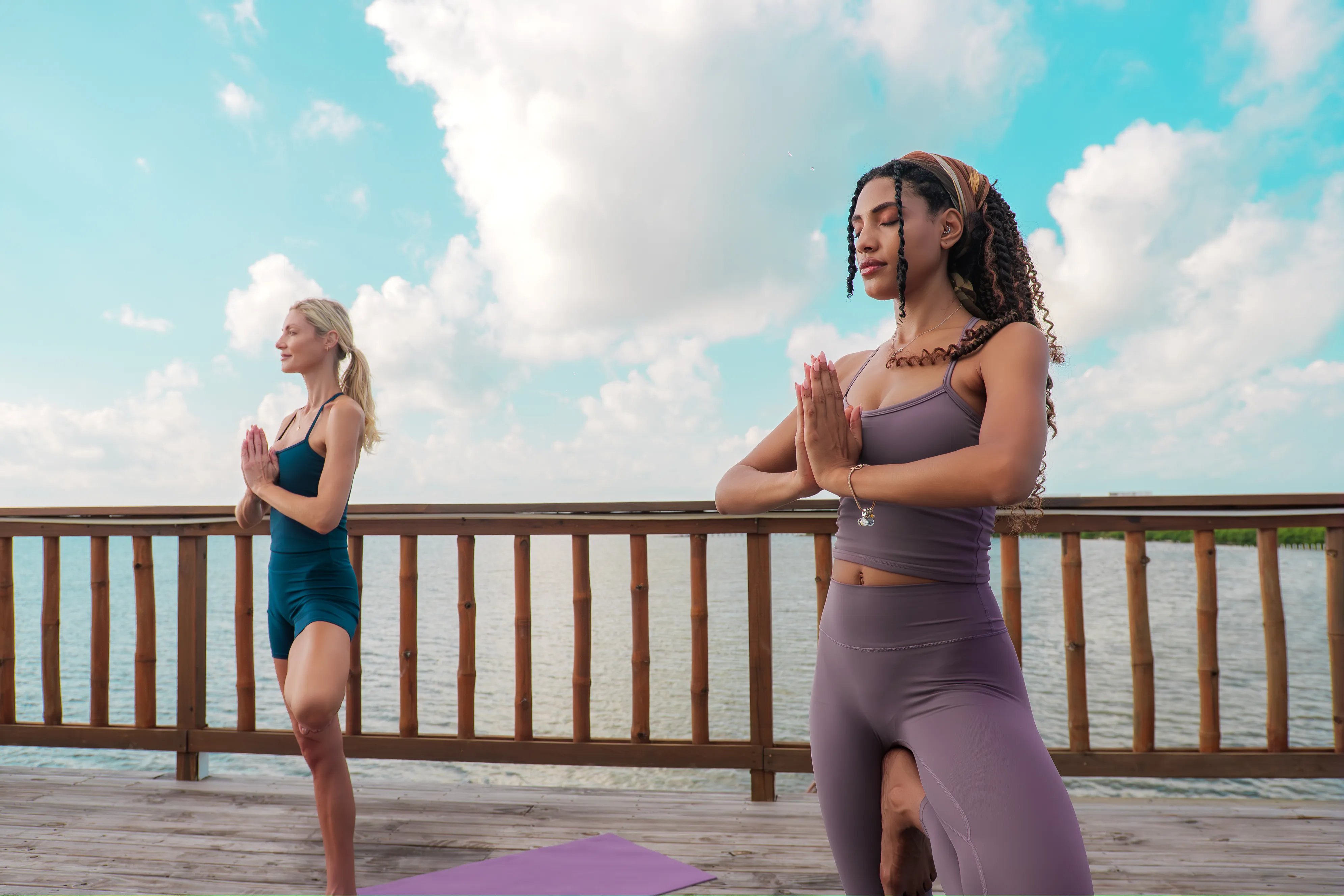 Two women practicing yoga in tree pose on a wooden deck by the water under a partly cloudy sky.