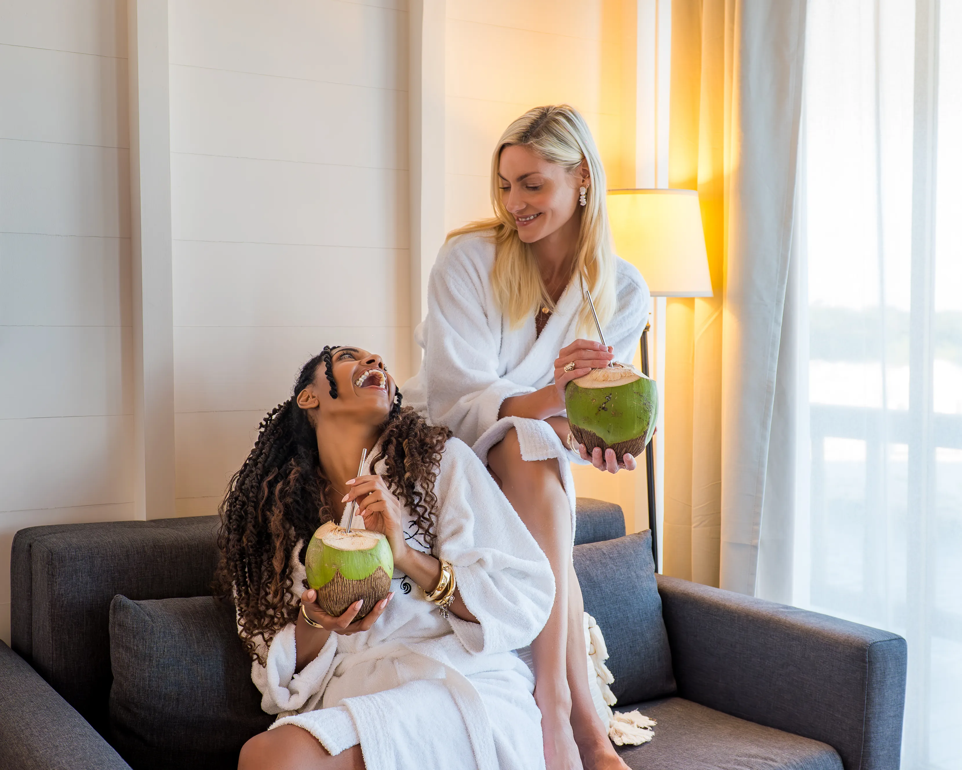 Two women wearing white bathrobes sitting on a couch, drinking fresh coconut water and smiling at each other.
