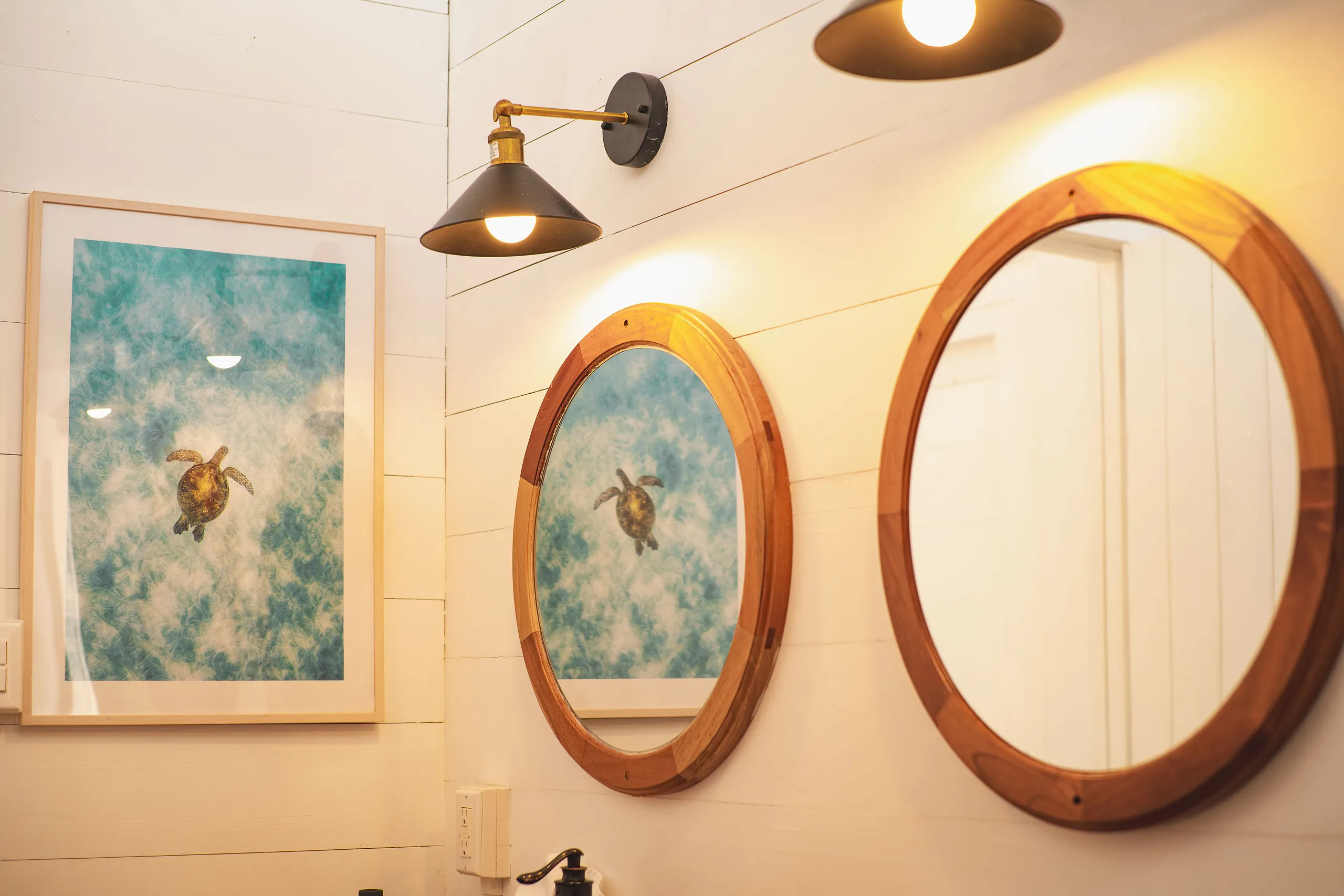 Bathroom wall with two round wooden mirrors reflecting a framed picture of a sea turtle underwater and a lit black and gold wall lamp.