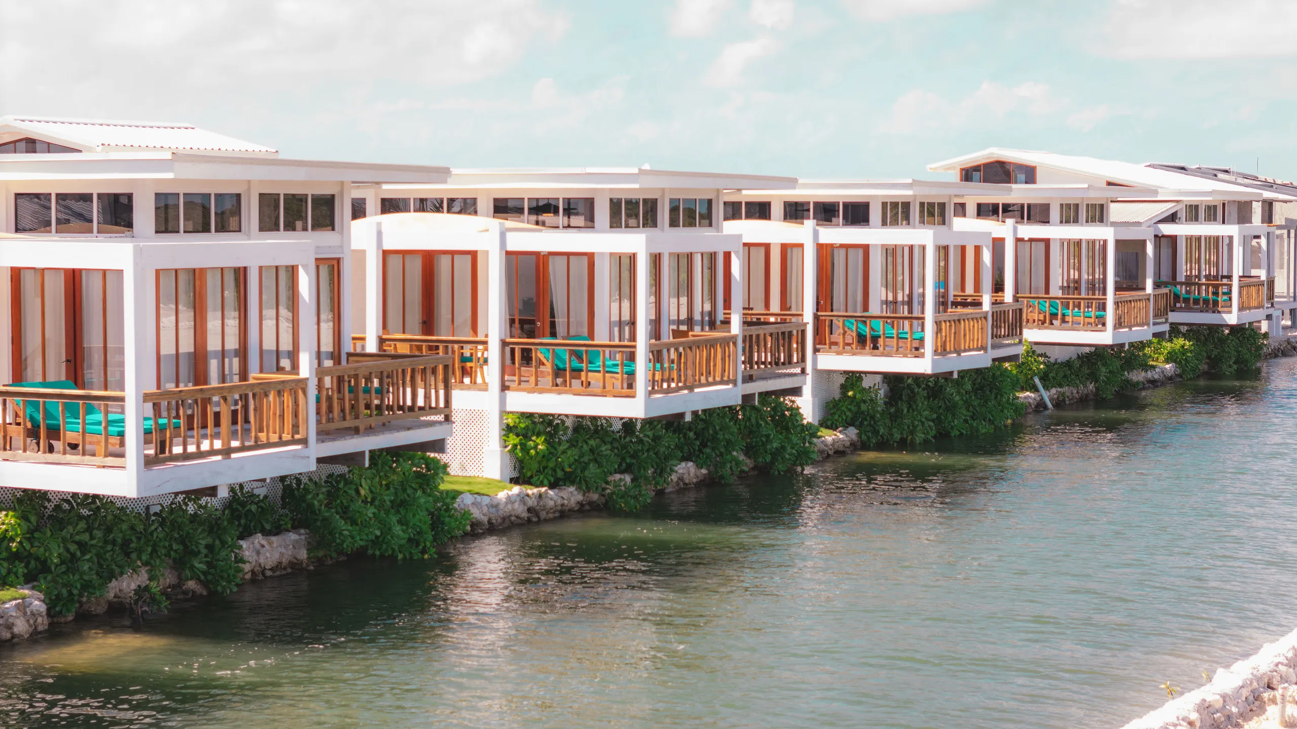Row of modern white waterfront villas with wooden balconies and teal lounge chairs overlooking calm water.