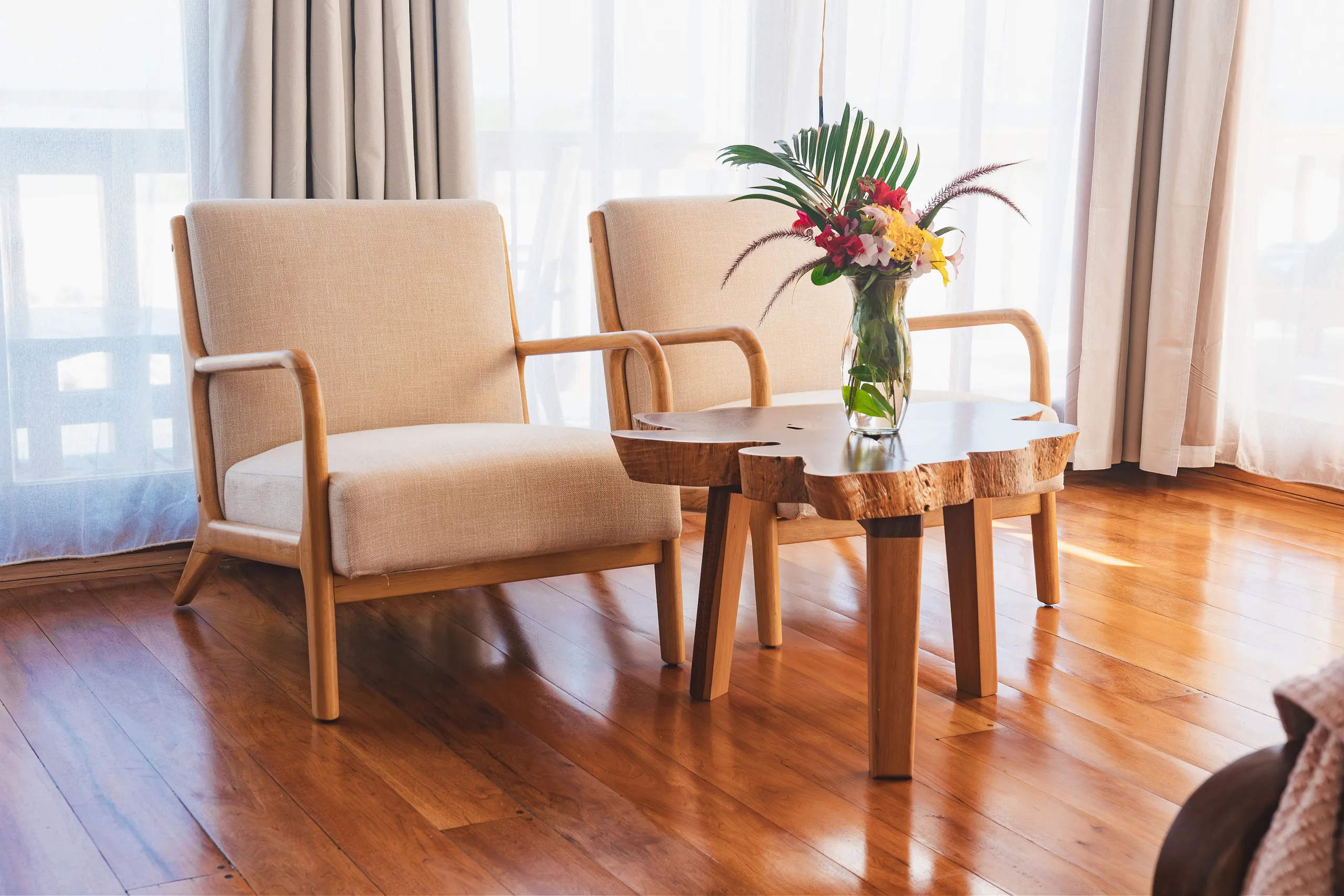 Two beige armchairs with wooden frames next to a natural edge wooden coffee table topped with a vase of colorful flowers in a sunlit room with wooden floors and white curtains.