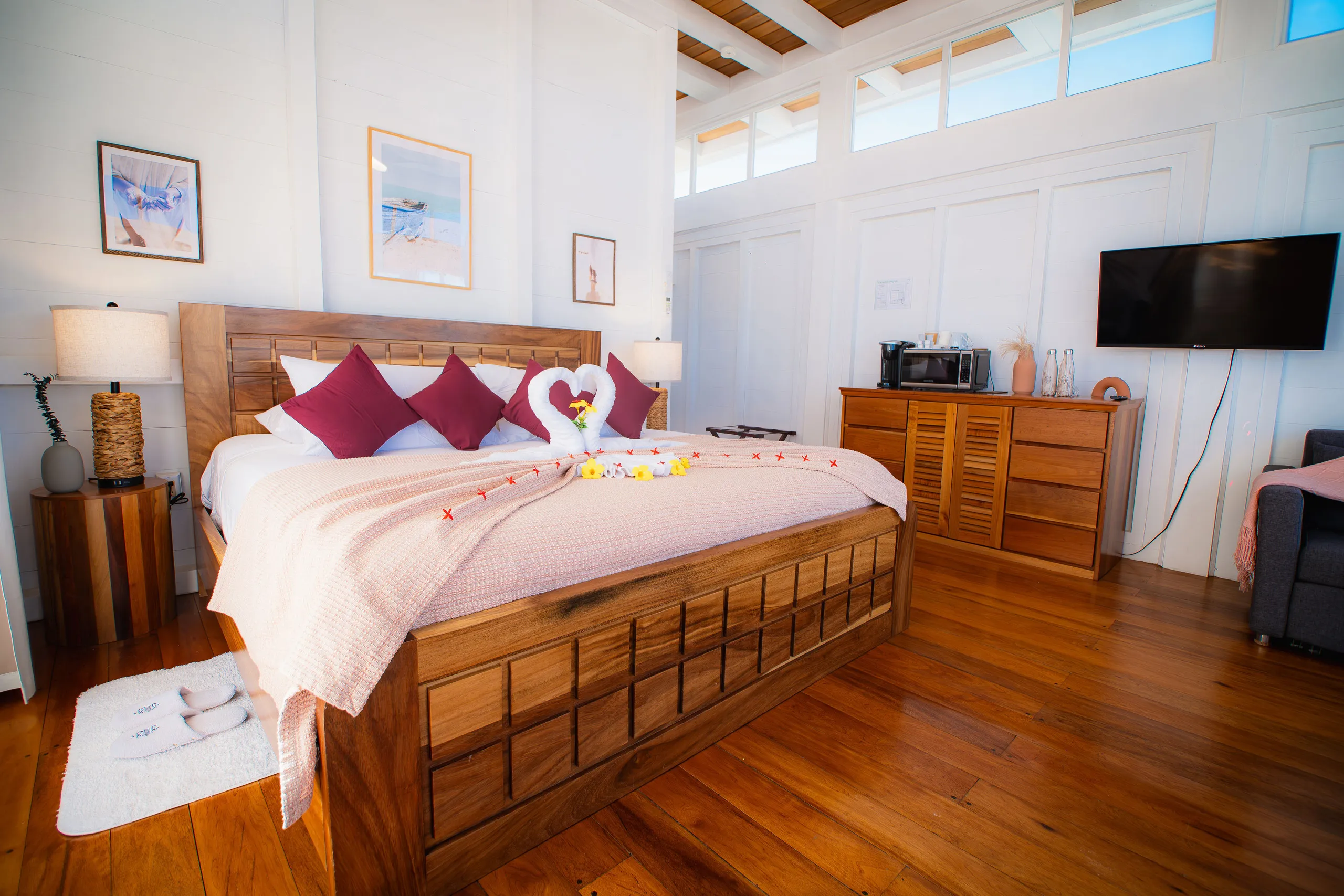 Bright bedroom with wooden bed frame, pink blanket, red pillows, towel swans with flowers, wooden dresser, and wall-mounted TV.