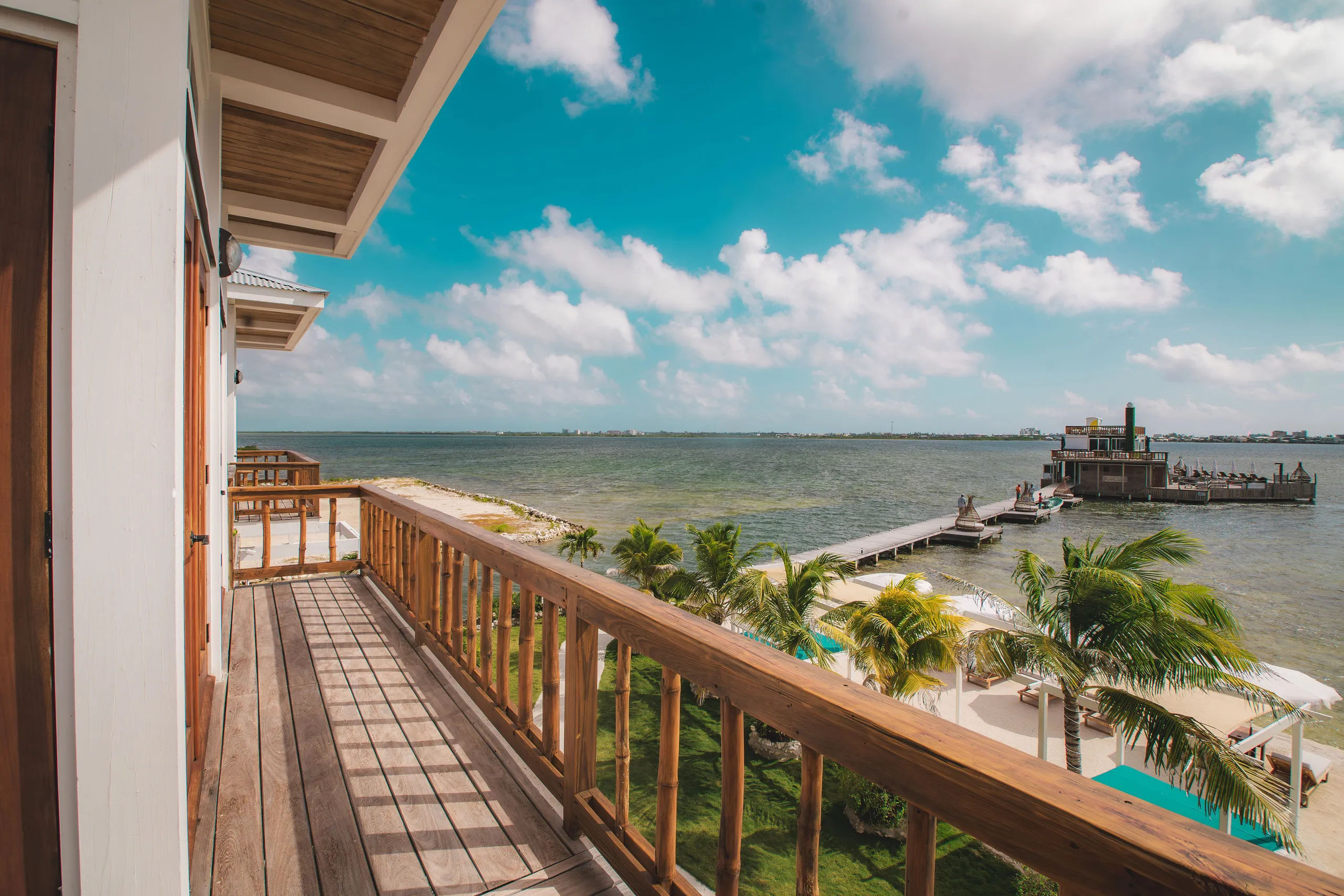 View from a wooden balcony overlooking palm trees, beach umbrellas, a pier extending into the water, and a partly cloudy blue sky.