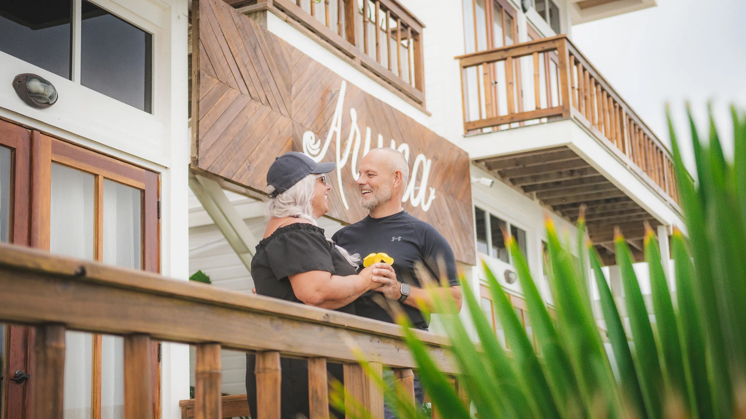 Smiling couple holding hands and standing on a wooden balcony in front of a building with a wooden sign that reads 'Arima'.
