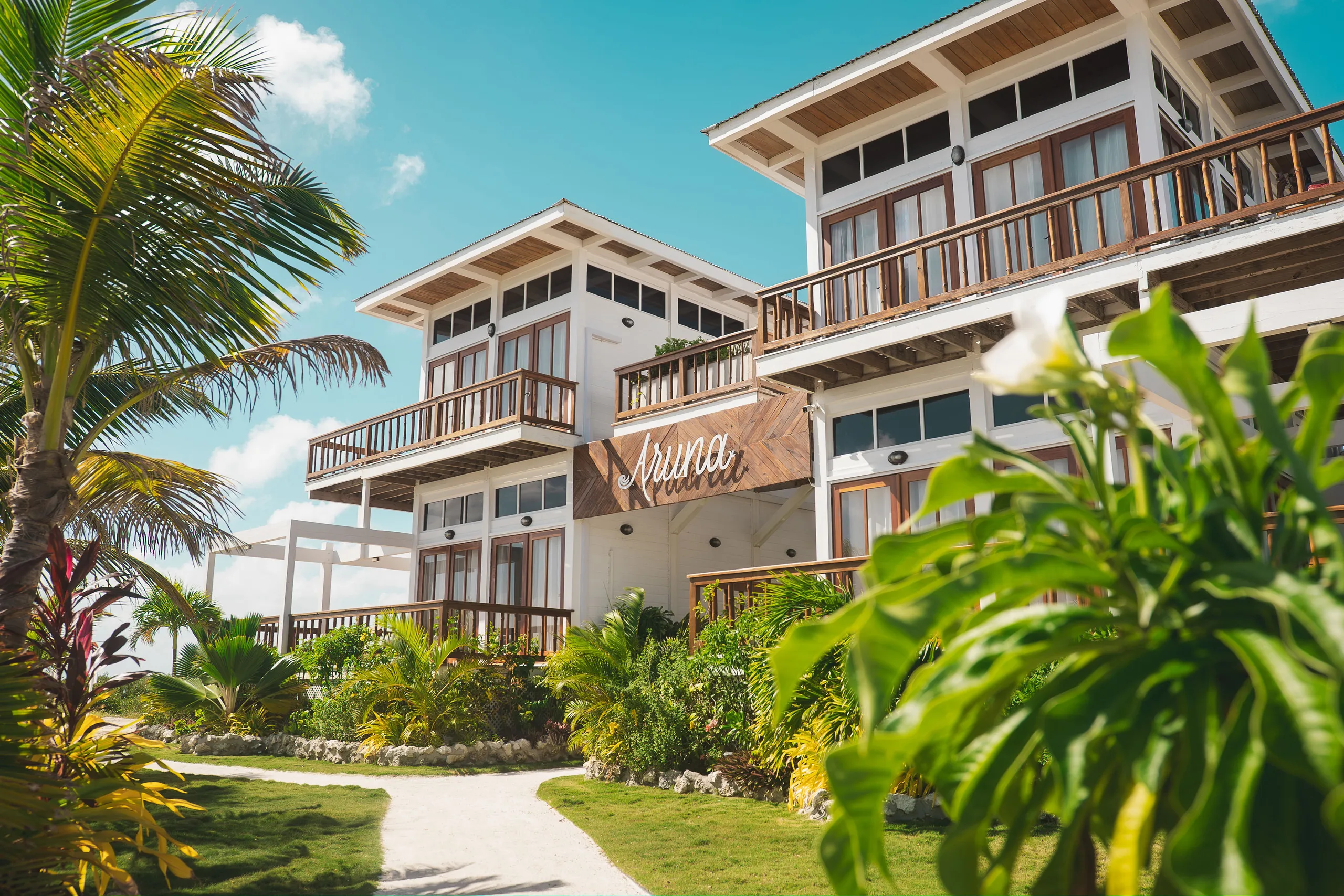 Modern tropical resort buildings with wooden balconies surrounded by lush green plants and palm trees under a clear blue sky.