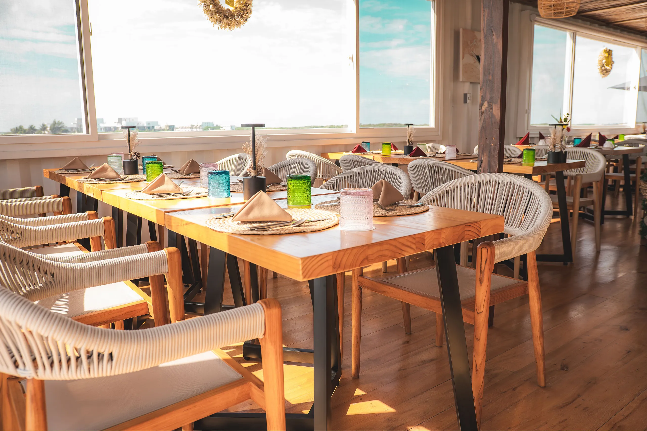 Bright restaurant interior with wooden tables set with colorful glasses and beige napkins, surrounded by woven and wooden chairs.