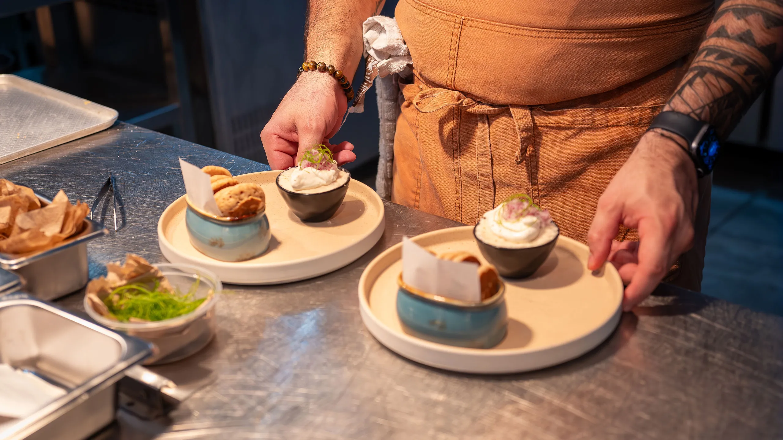Chef wearing a brown apron plating two dishes with cookies and a creamy dessert in small bowls on beige plates.
