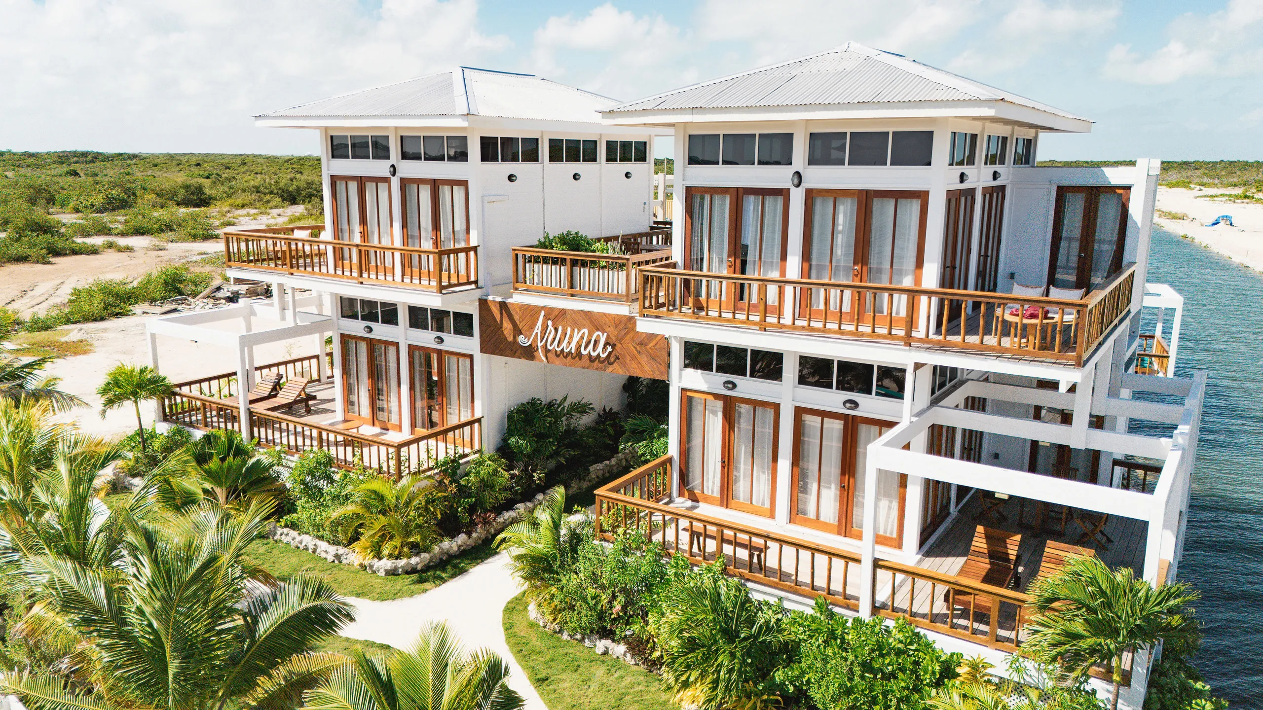 Modern white two-story building with wooden balconies surrounded by palm trees and greenery near a canal.