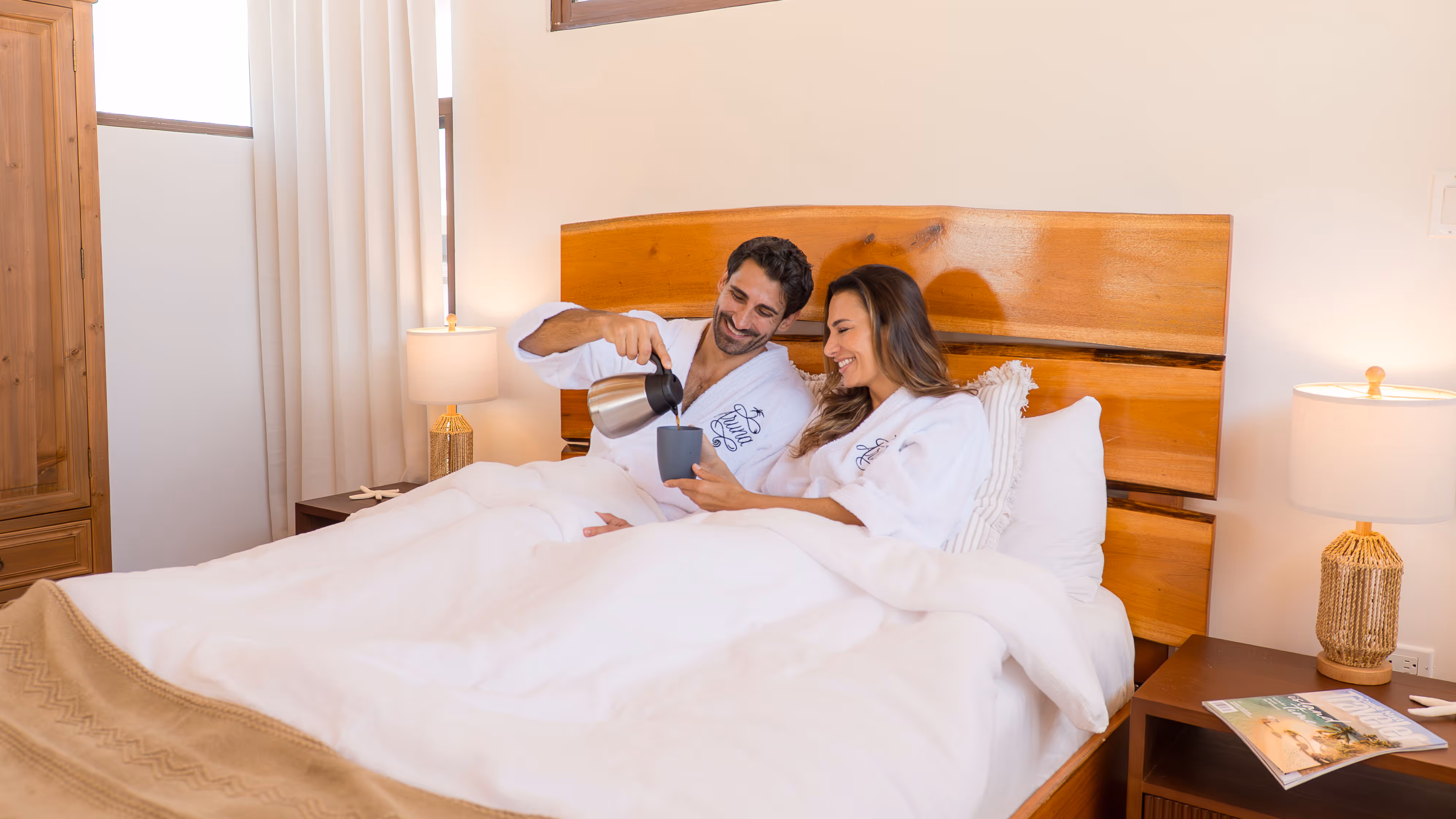 Smiling couple in white robes sitting in bed while man pours coffee into a mug held by the woman.