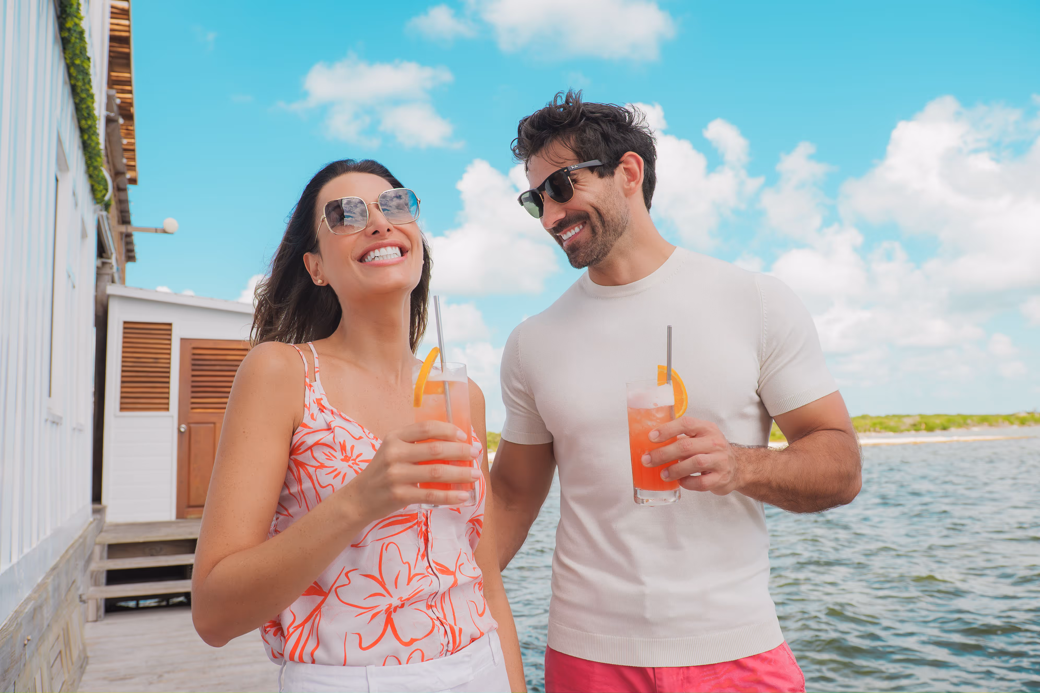 Smiling man and woman wearing sunglasses, holding orange drinks with straws, standing on a wooden dock by the water under a blue sky with clouds.
