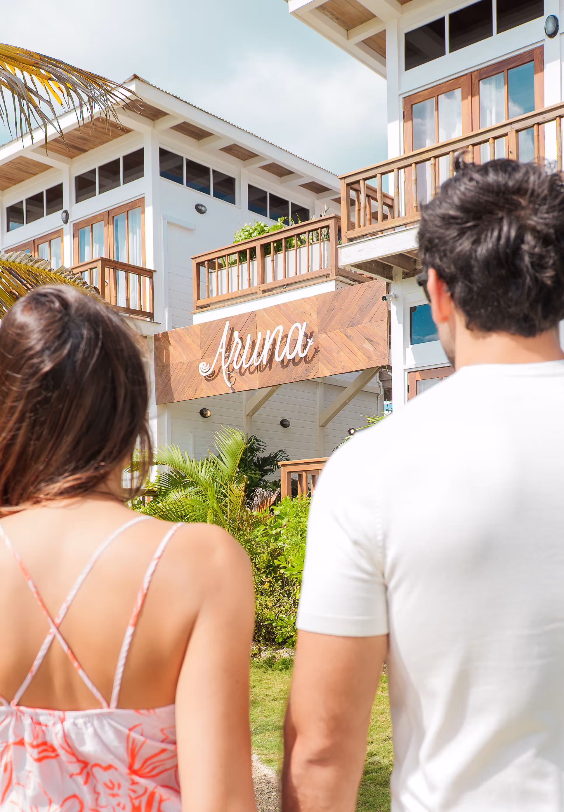 Couple holding hands, facing away towards a white wooden building with balconies and a wooden sign reading 'Aruna'.