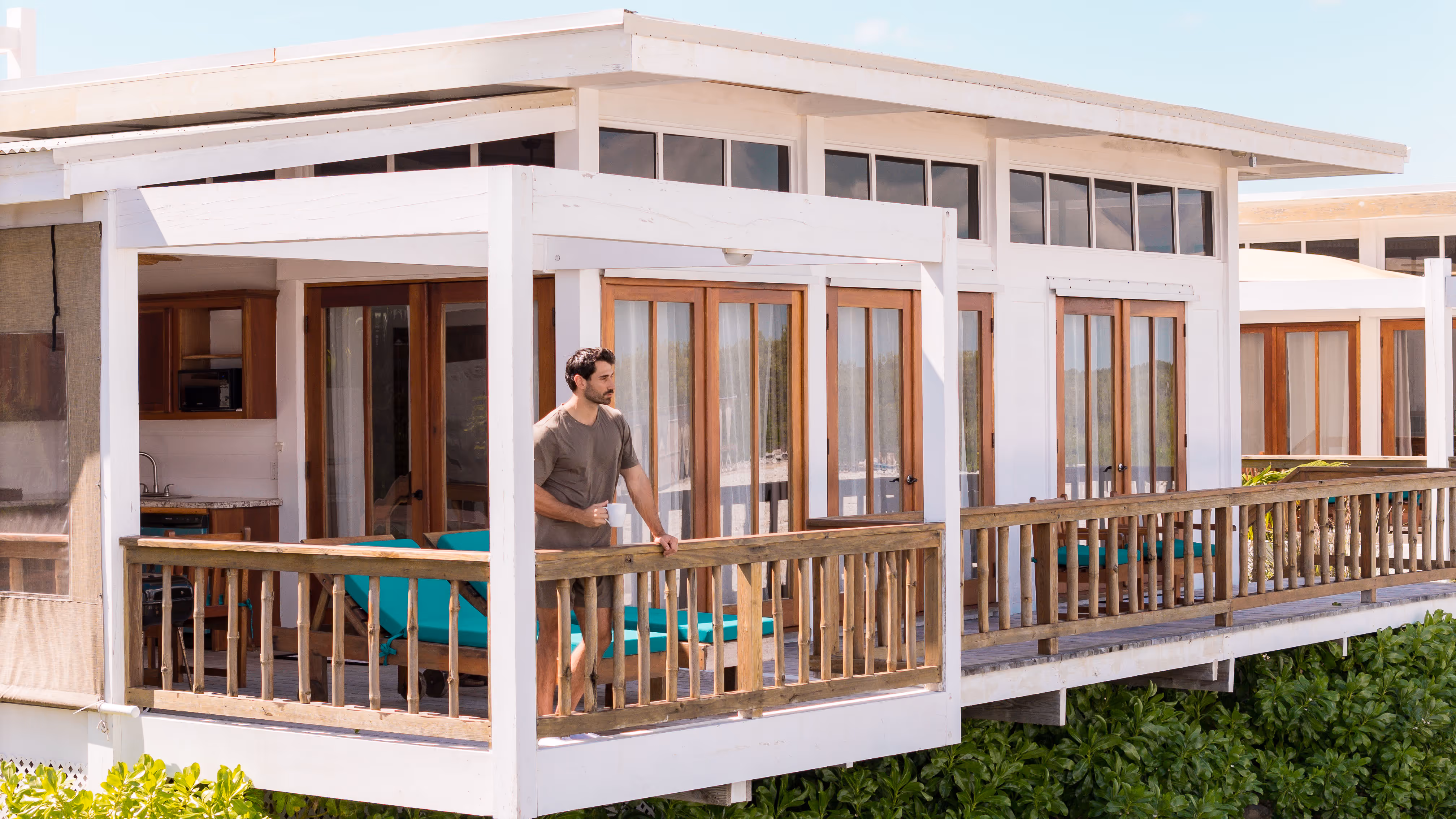 Man in casual clothes holding a cup standing on a wooden balcony of a white beach house with turquoise lounge chairs.