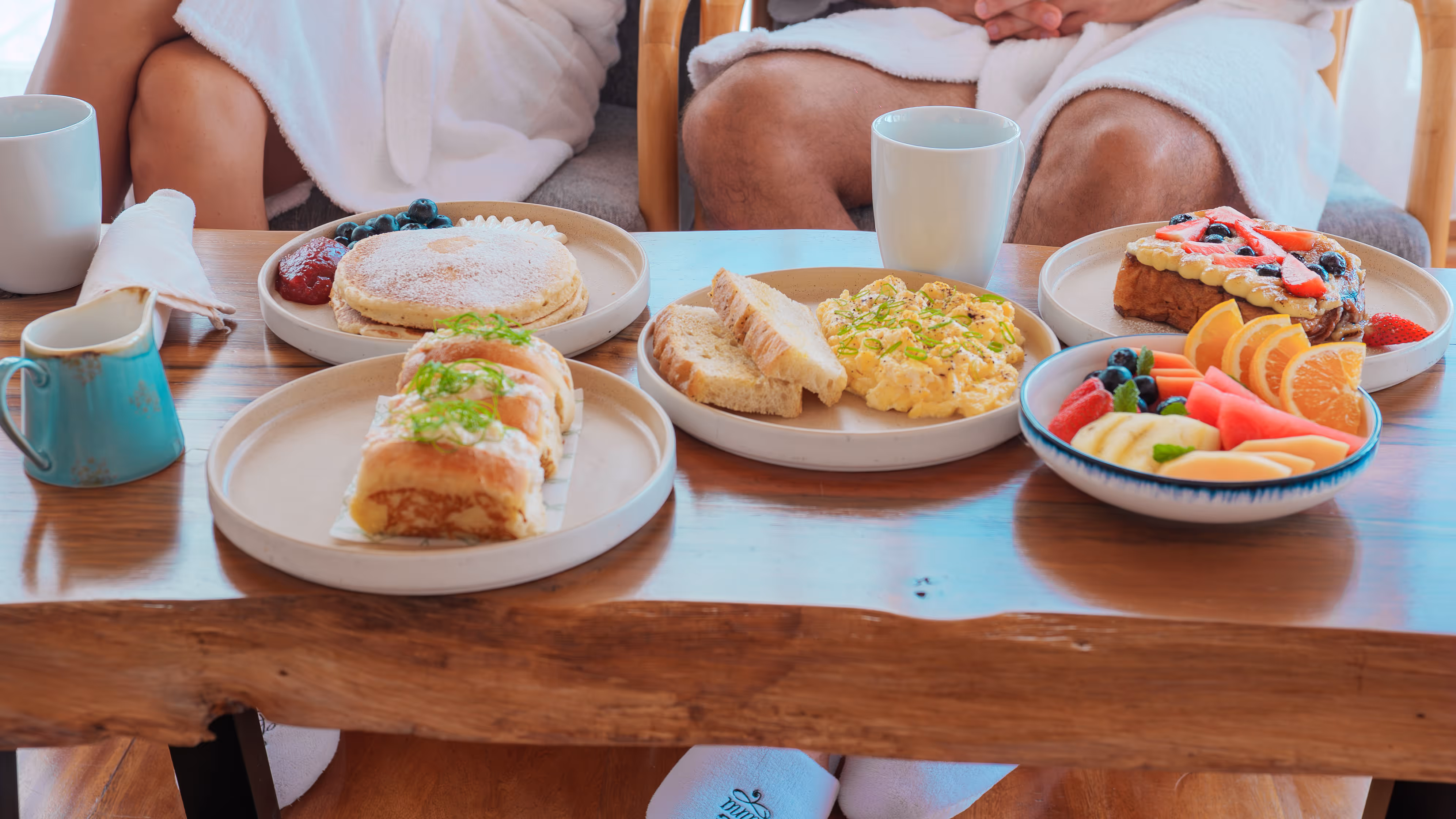 Breakfast spread on a wooden table with pancakes, scrambled eggs with toast, fruit salad, buns, and two white mugs.