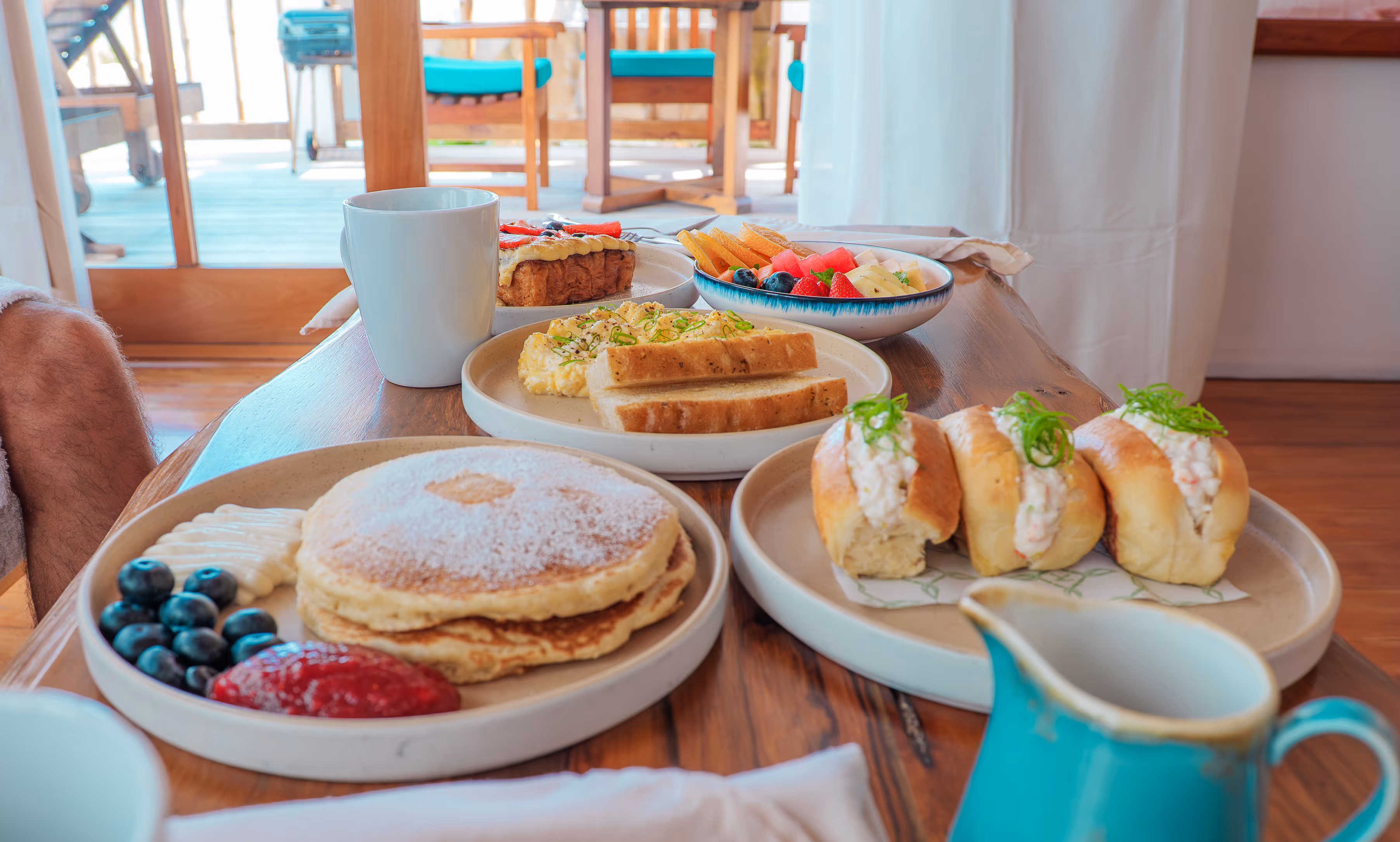 Breakfast spread on a wooden table including pancakes with blueberries and strawberry jam, scrambled eggs with toast, fruit bowl, shrimp rolls, a cup, and a turquoise pitcher.